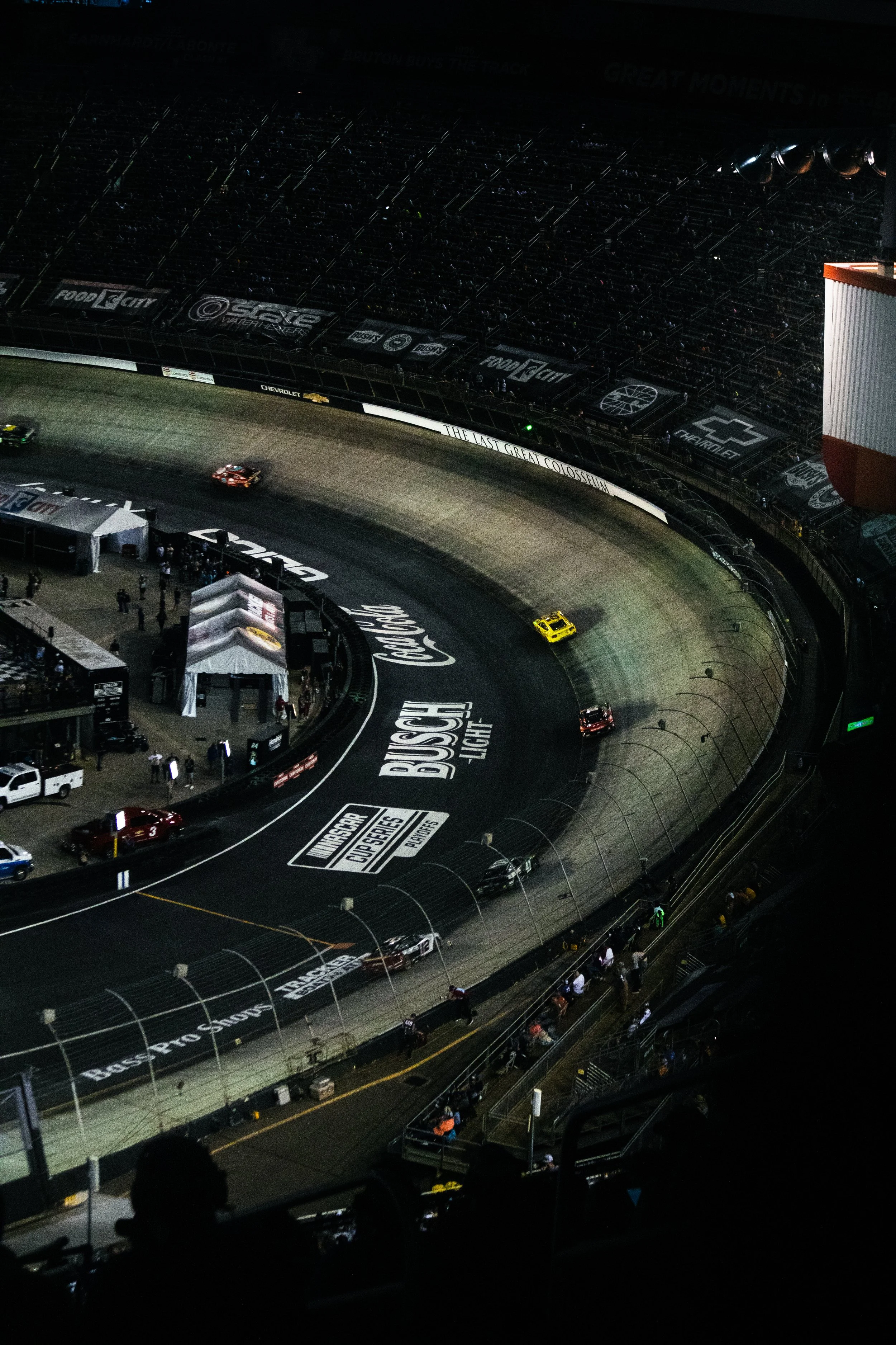 Nighttime view of a race track with several cars on the track and empty grandstand seats. The track has various sponsor logos and banners, including Ford, Chevrolet, and Busch Light.