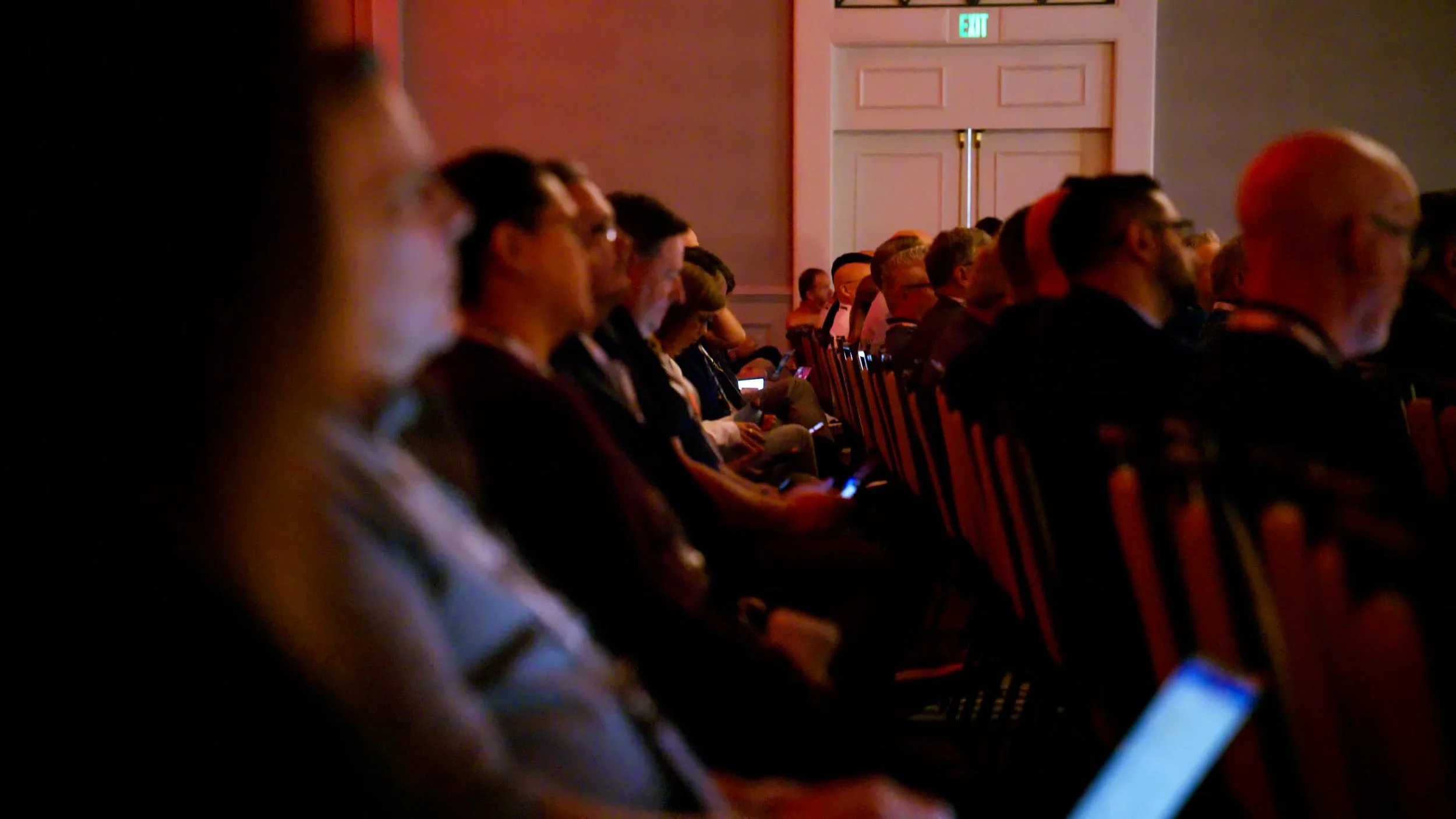 People seated in rows at a conference or event, some looking at their phones, in a dimly lit room with a pink wall and an exit sign