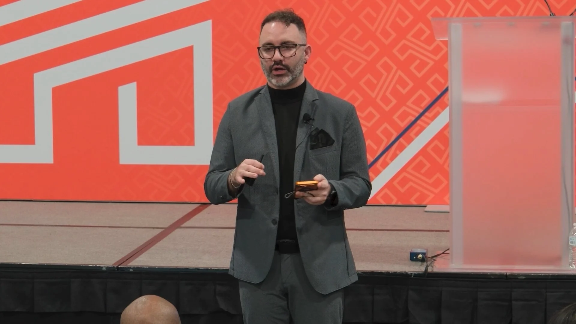 A man with glasses, a beard, and short hair dressed in a gray suit jacket, black turtleneck, and black pants, standing on a stage with a large orange and white geometric-patterned backdrop, holding a smartphone in his right hand, and speaking for an 