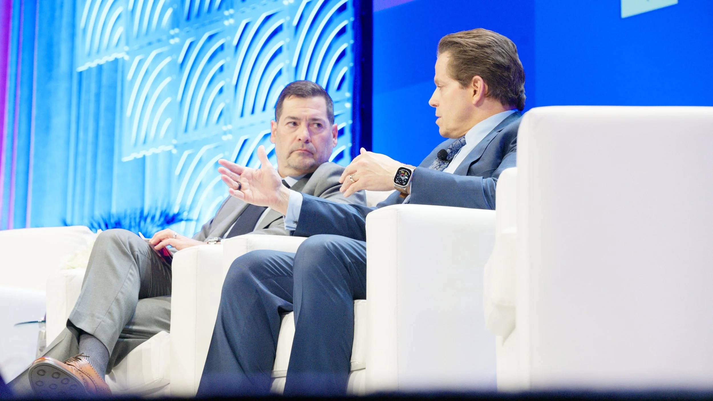 Two men are sitting on a stage during a panel discussion or conference, engaged in conversation, with a decorative blue backdrop behind them.