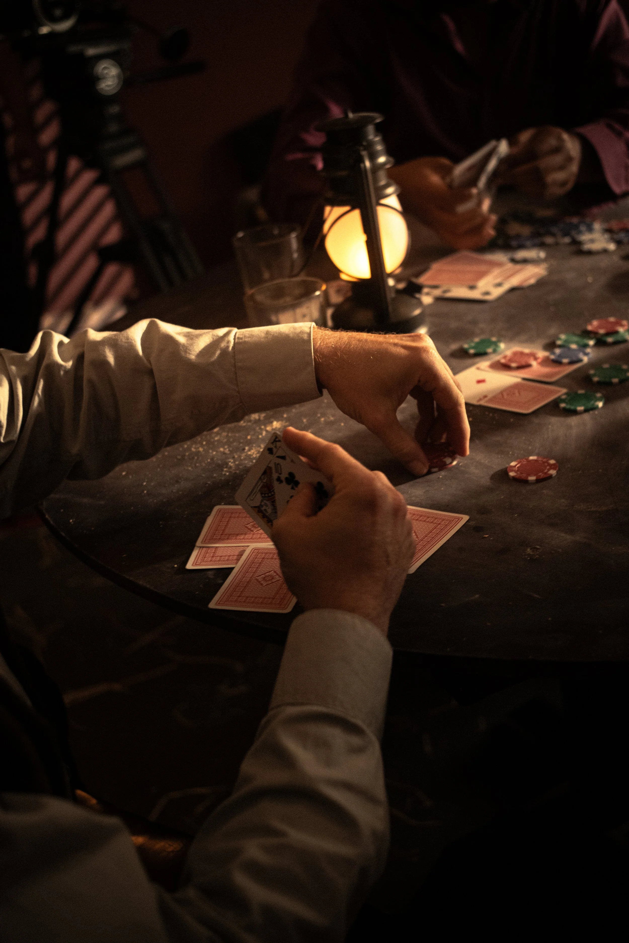 People playing poker at a dimly lit table, with poker chips and playing cards, and a lantern providing a warm glow.