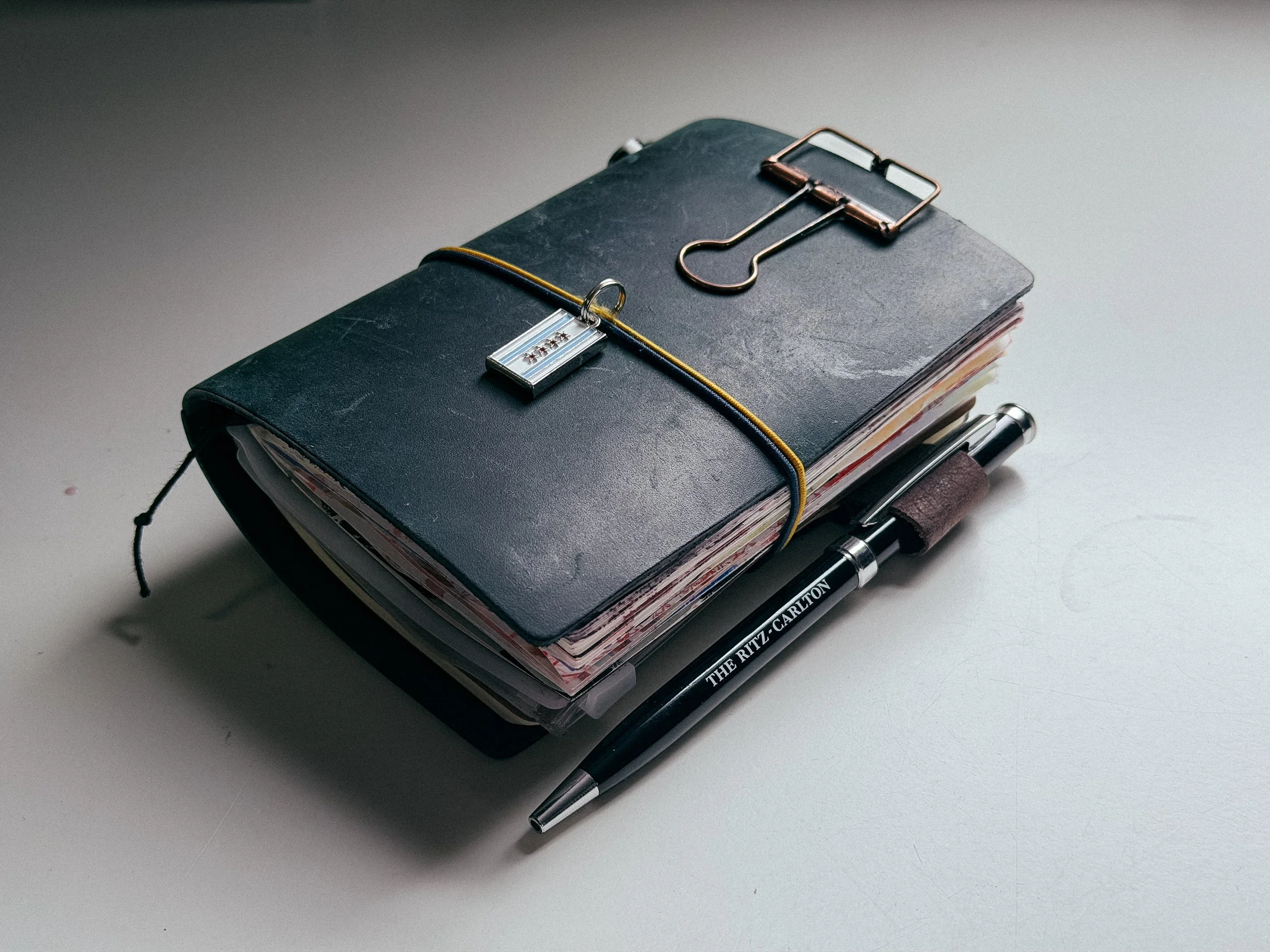 A black planner with a yellow and black elastic band, a small pen, and a metal clip with a decorative tag on top. A thick black marker is placed next to the planner, on a light-colored surface.