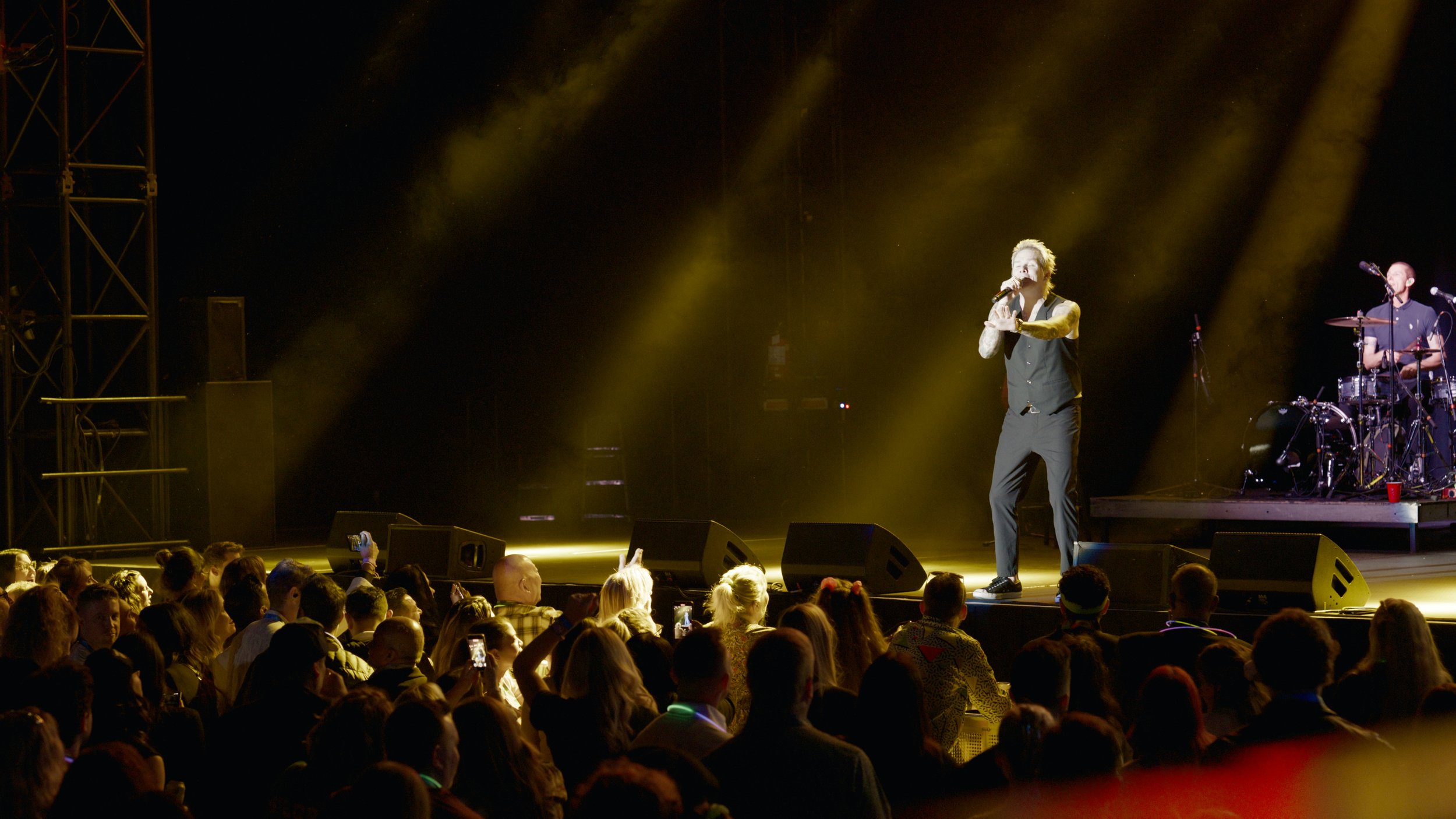 A male performer stands on stage singing into a microphone with a band member playing drums in the background, illuminated by yellow stage lights, in front of an audience.