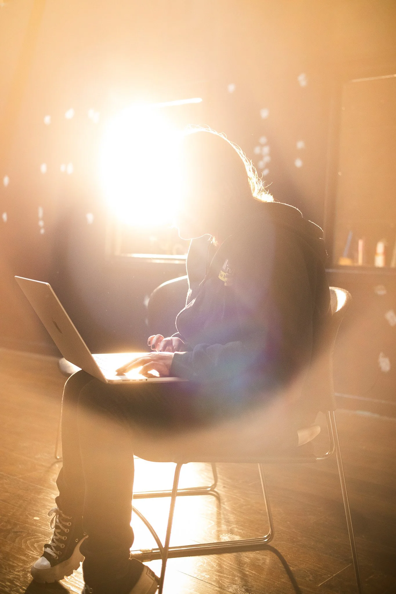 A woman sitting at a table using a laptop, with bright sunlight shining behind her, creating a silhouette effect.