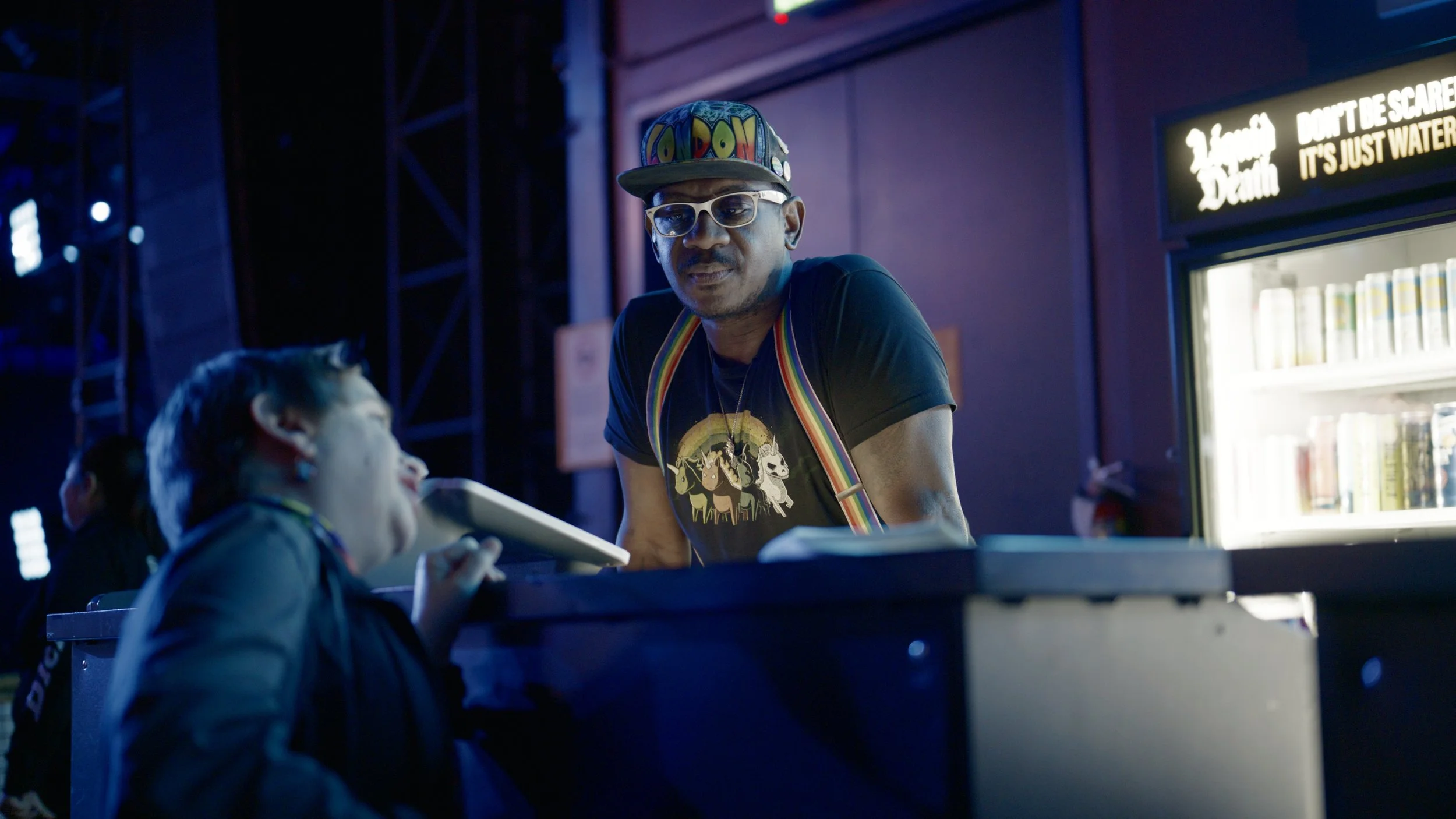 A man wearing glasses, a colorful cap, and a black t-shirt with a rainbow and horses graphic, is standing at a counter interacting with a woman in a dark jacket at night. There is a vending machine with drinks and a sign that says 'Don't be scared, i