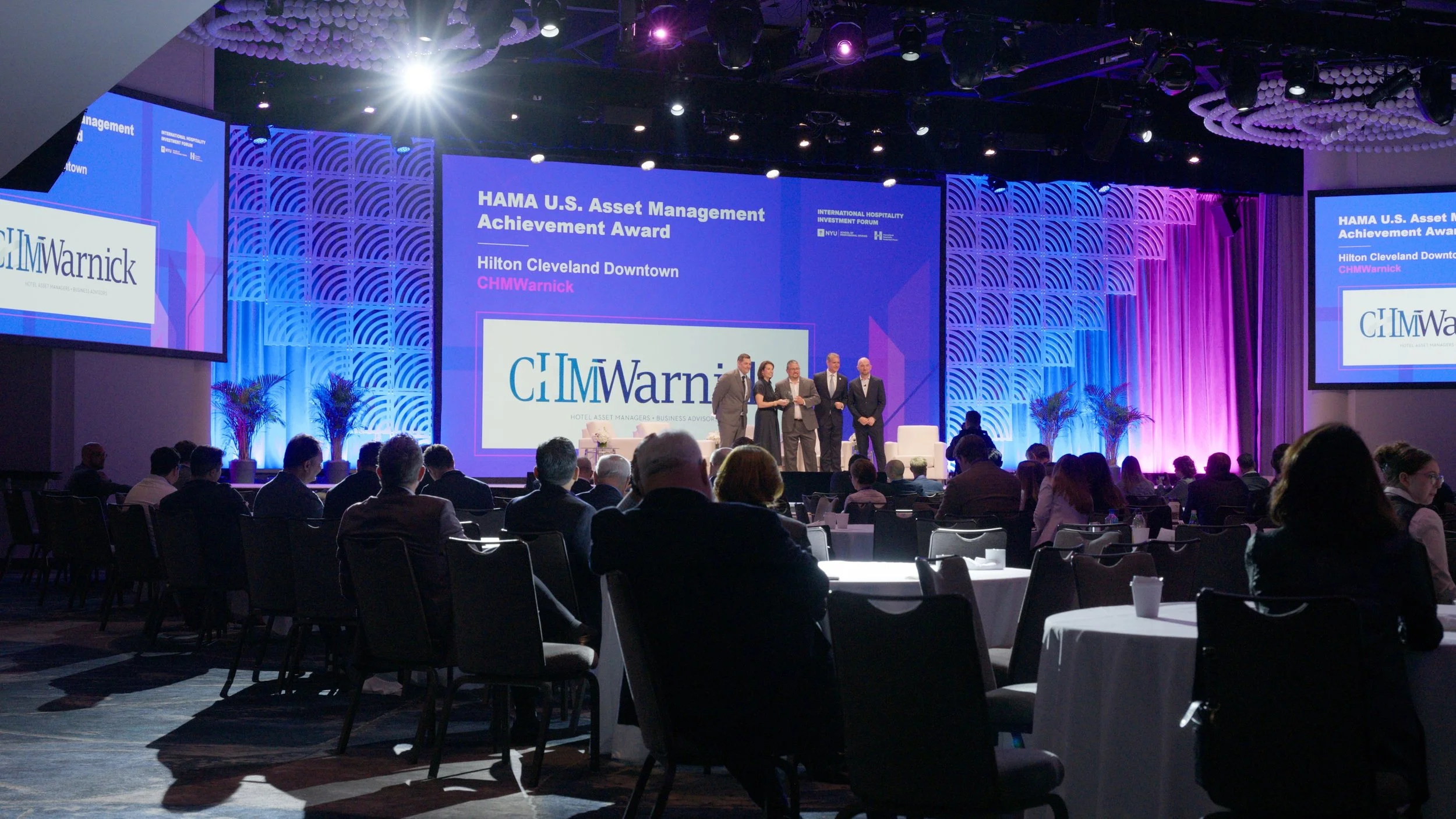 A stage at an awards event with large screens displaying text about the 'HAMA U.S. Asset Management Achievement Award' for Hilton Cleveland Downtown CHMWarnick. Four people are standing on stage, speaking to an audience seated at round tables in a co