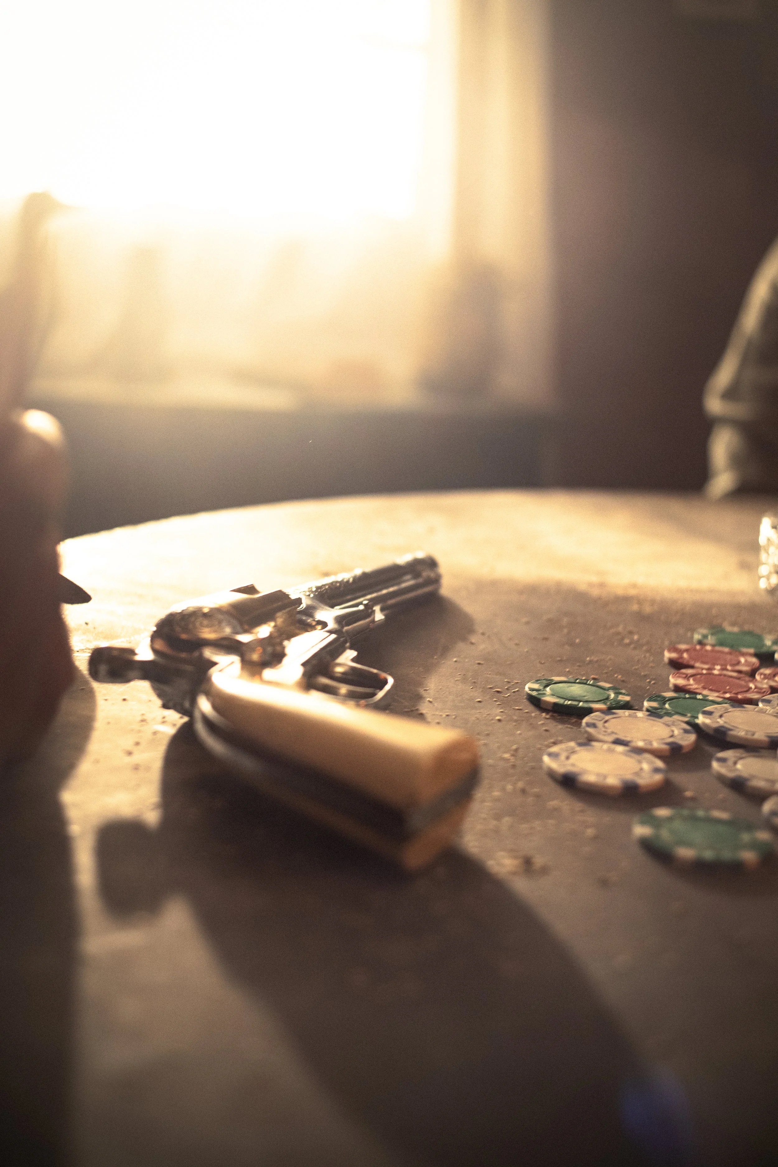 Close-up of poker chips and a gun on a wooden table, with sunlight streaming through a window in the background.