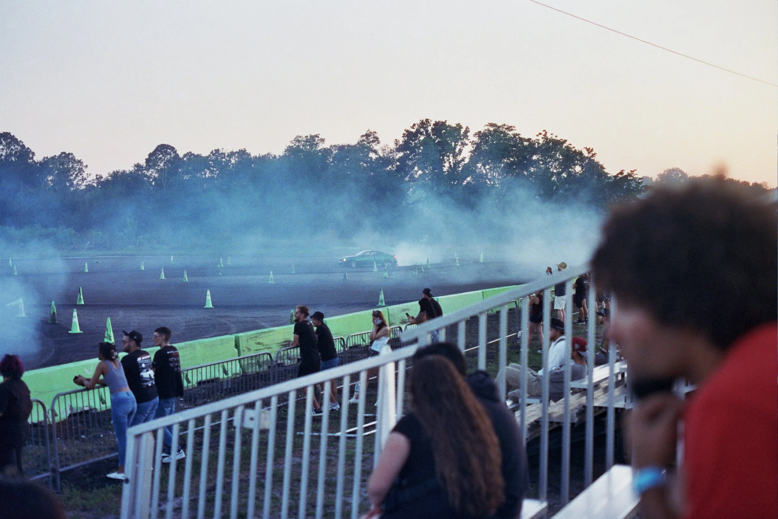 People standing behind a white fence at a car drifting event during the evening, with smoke billowing from the tires and a car on the track.