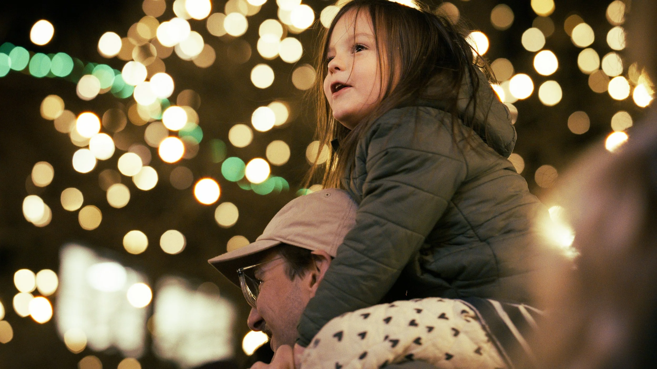 A young girl sitting on a person's shoulders, surrounded by blurred holiday lights at night.