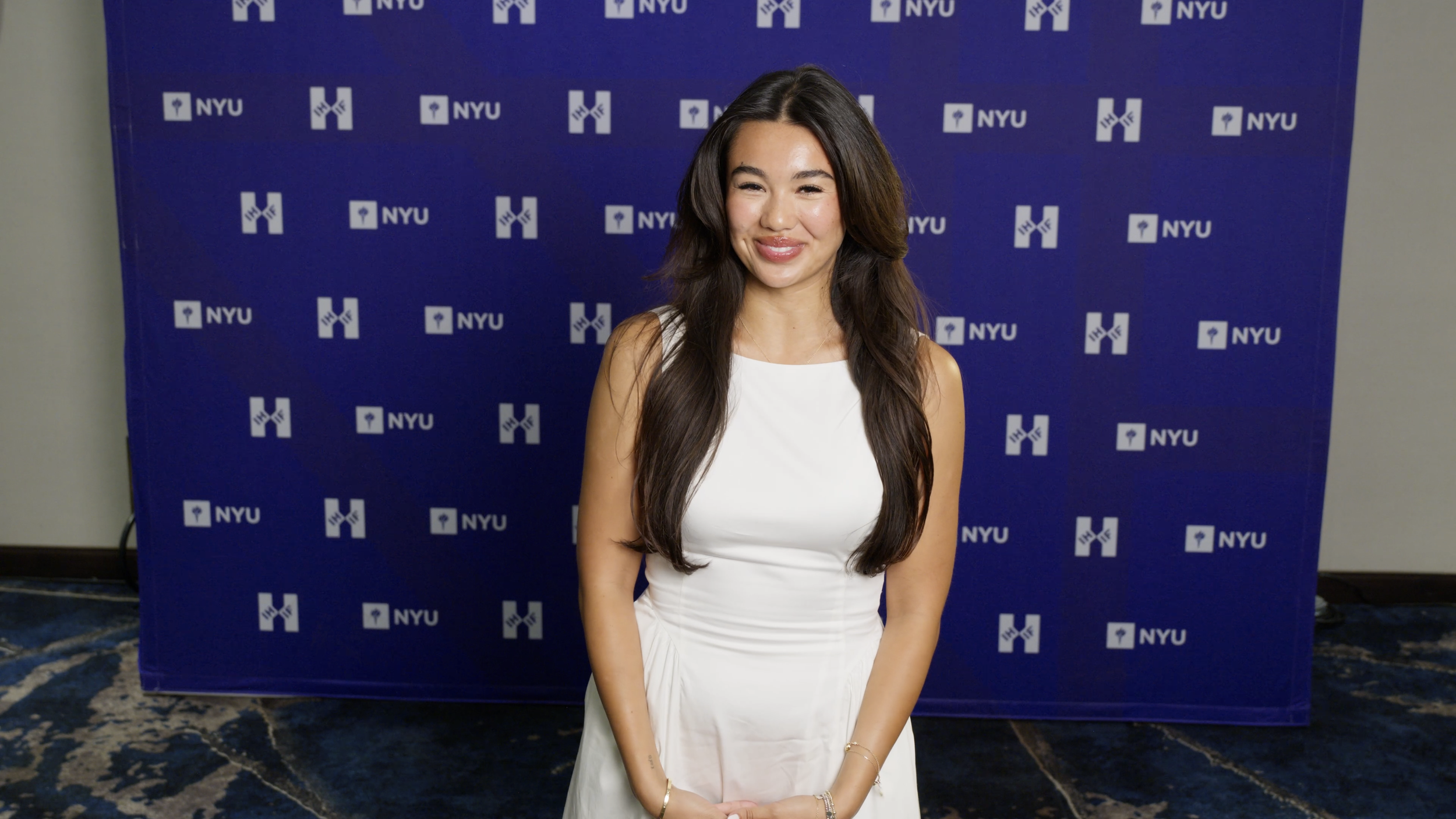 Young woman with long brown hair wearing a white dress, standing in front of a blue backdrop with the NYU logo.
