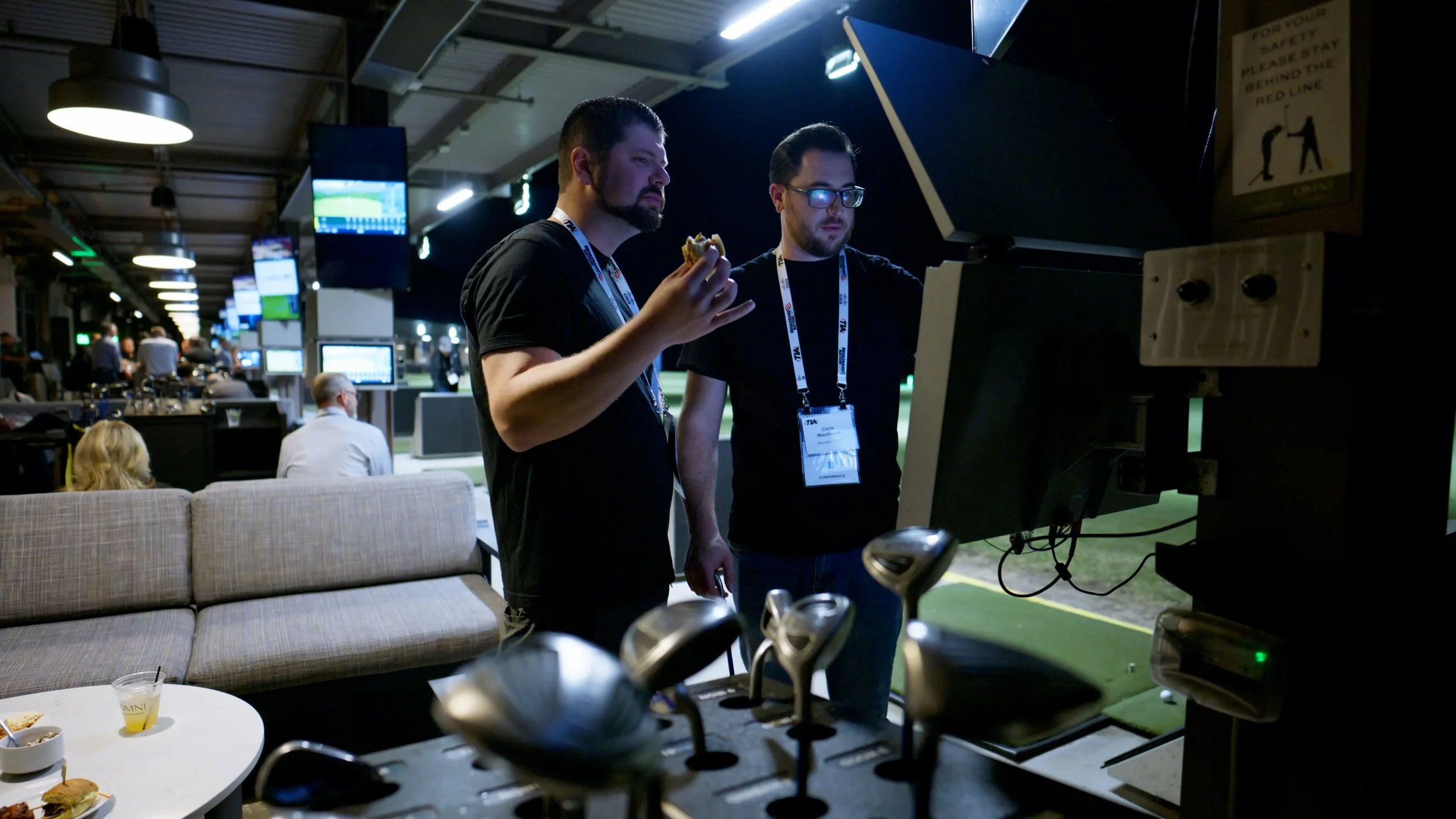 Two men working at a sports event using a computer, with golf clubs and snacks in the foreground, and a golf game on screens in the background.