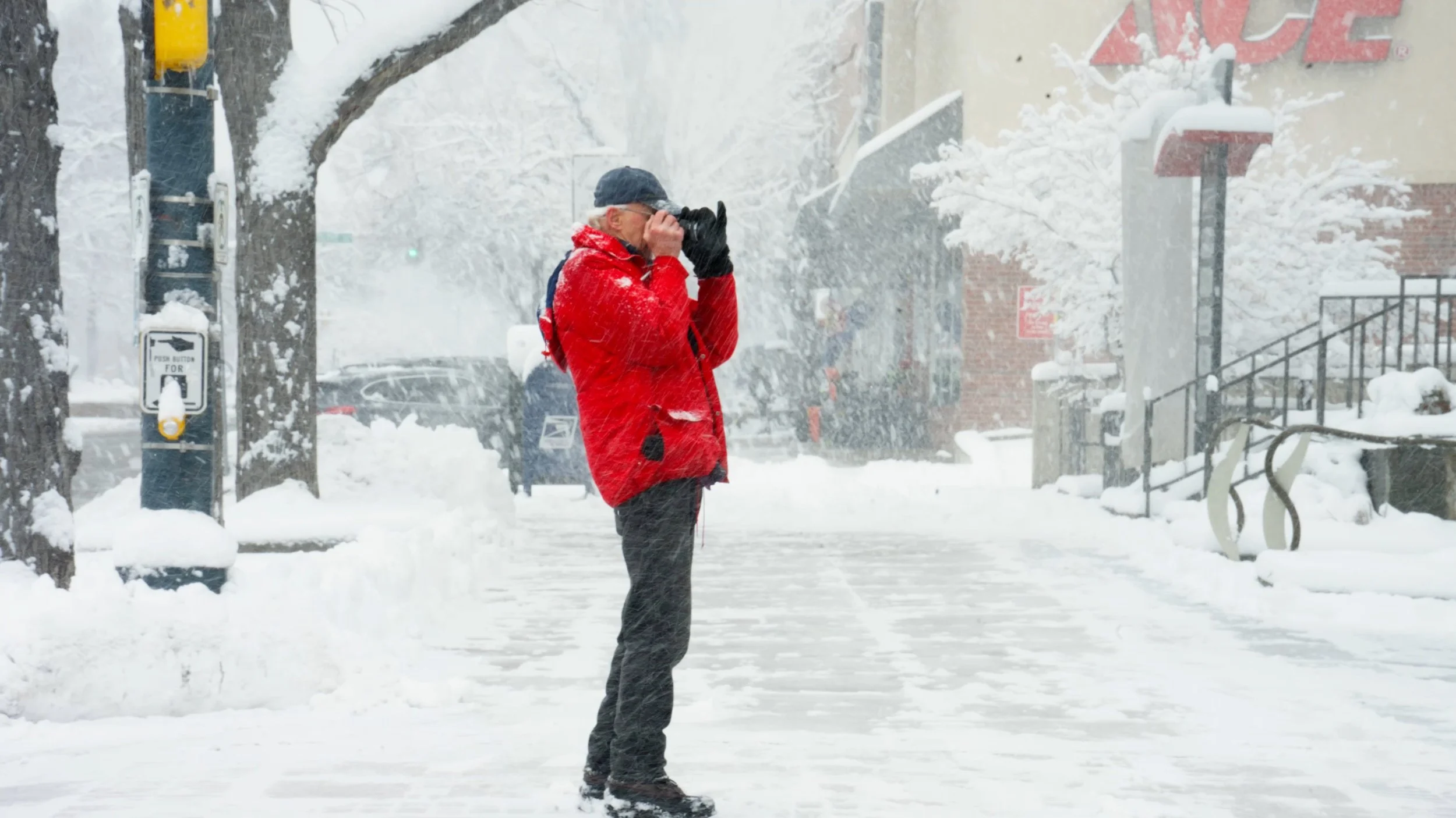 A person dressed in a red winter jacket and black gloves standing on a snow-covered sidewalk during a snowstorm, using a camera or binoculars to look into the distance.
