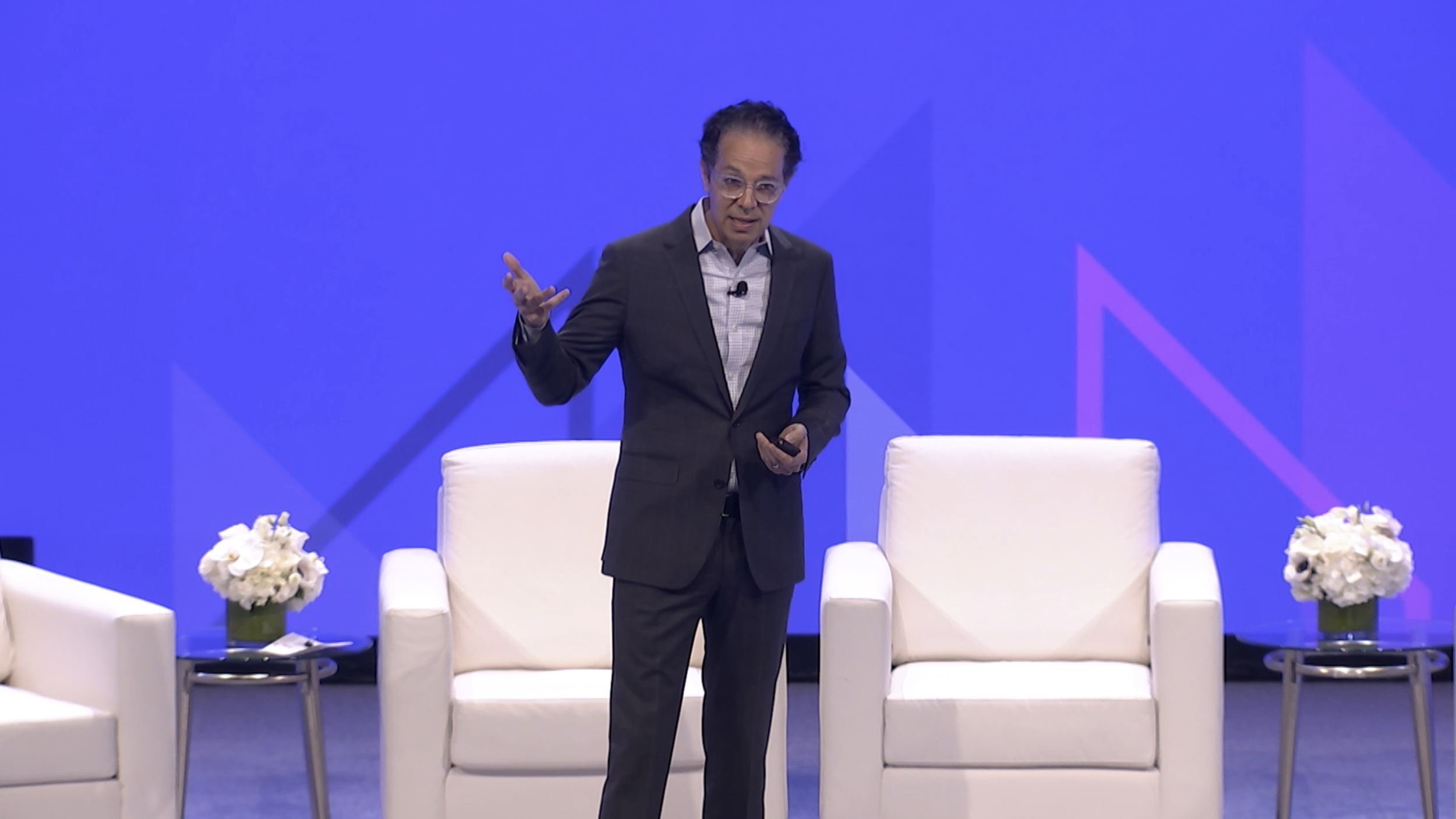 A man in a dark suit gives a presentation on a stage with a blue background and purple geometric designs, sitting chairs and flower arrangements are visible.