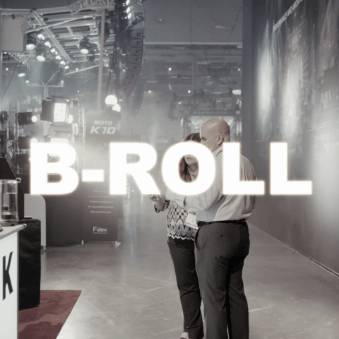 Two people sharing a moment at a trade show or expo, with a large illuminated sign reading 'B-ROLL' in the foreground.