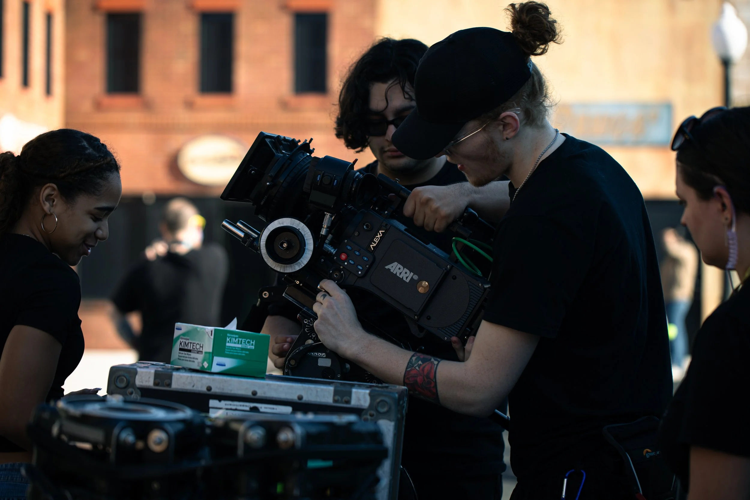 Group of people working with filming equipment outdoors, with a brick building in the background.