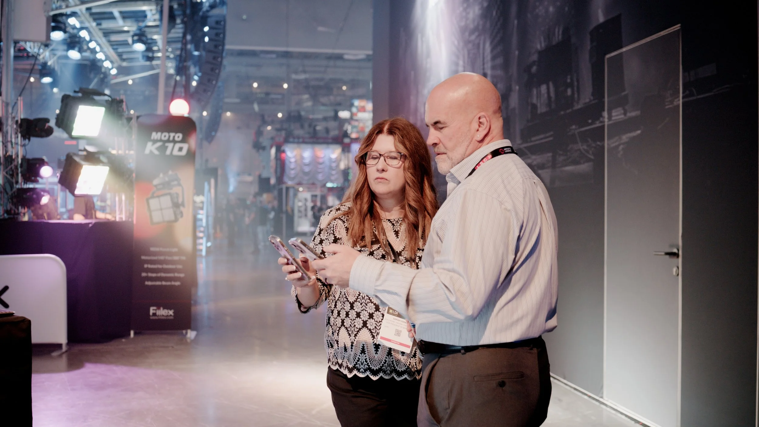 A woman with red hair and glasses and a man with a bald head and beard looking at a smartphone together at an indoor event or conference, with a background of stage lights and a promotional banner.