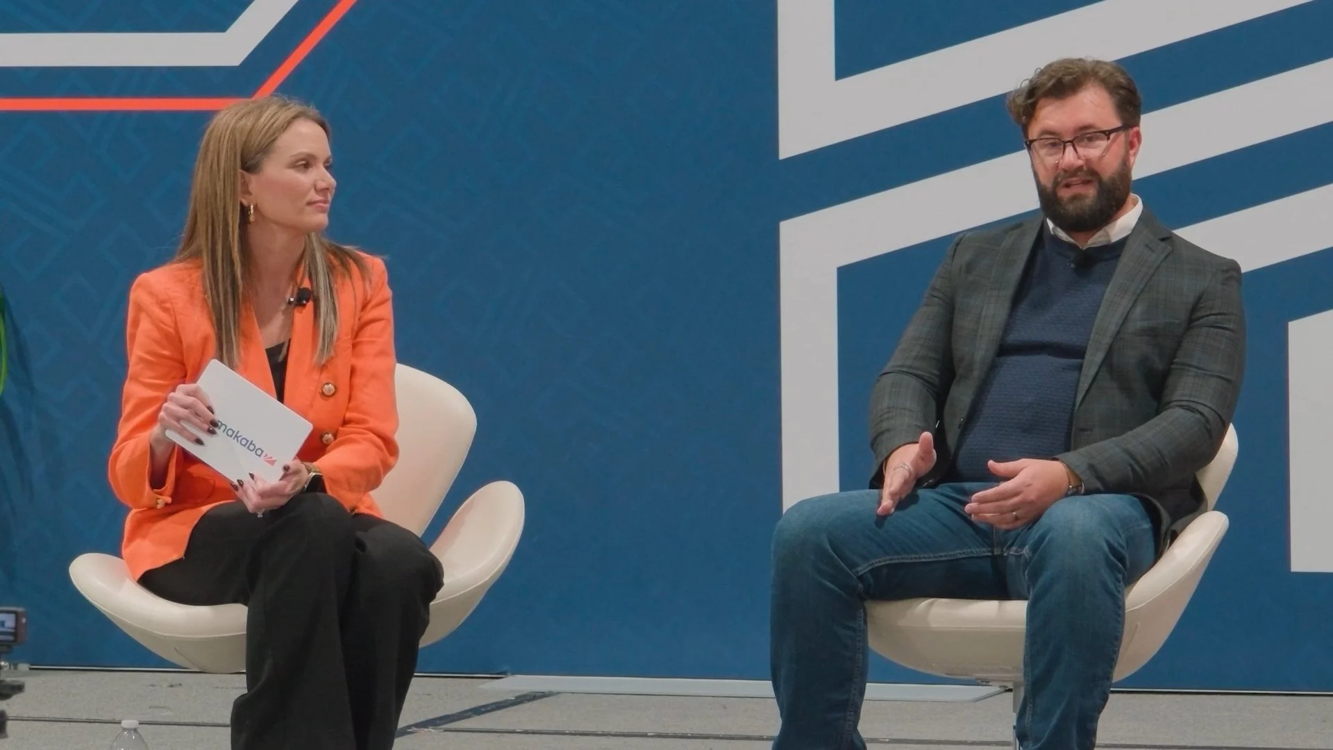 Two people seated on stage during a panel discussion. The woman on the left has long blond hair, wearing an orange blazer and holding a notecard. The man on the right has a beard and glasses, wearing a checkered blazer with a blue sweater and jeans.