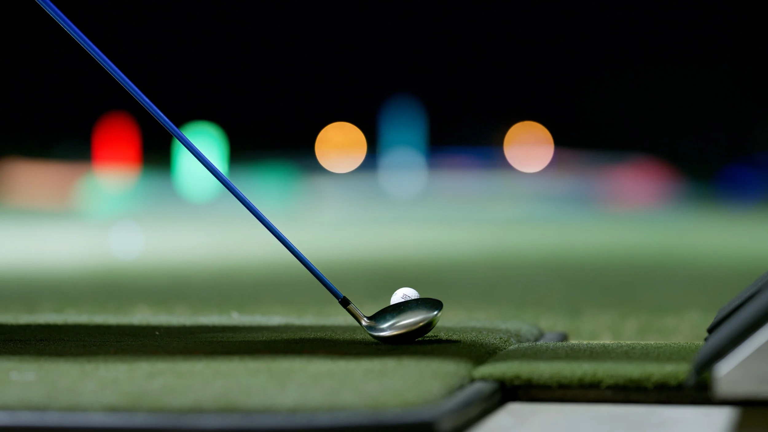 Close-up of a golf club striking a golf ball on a putting green, with colored lights blurred in the background.