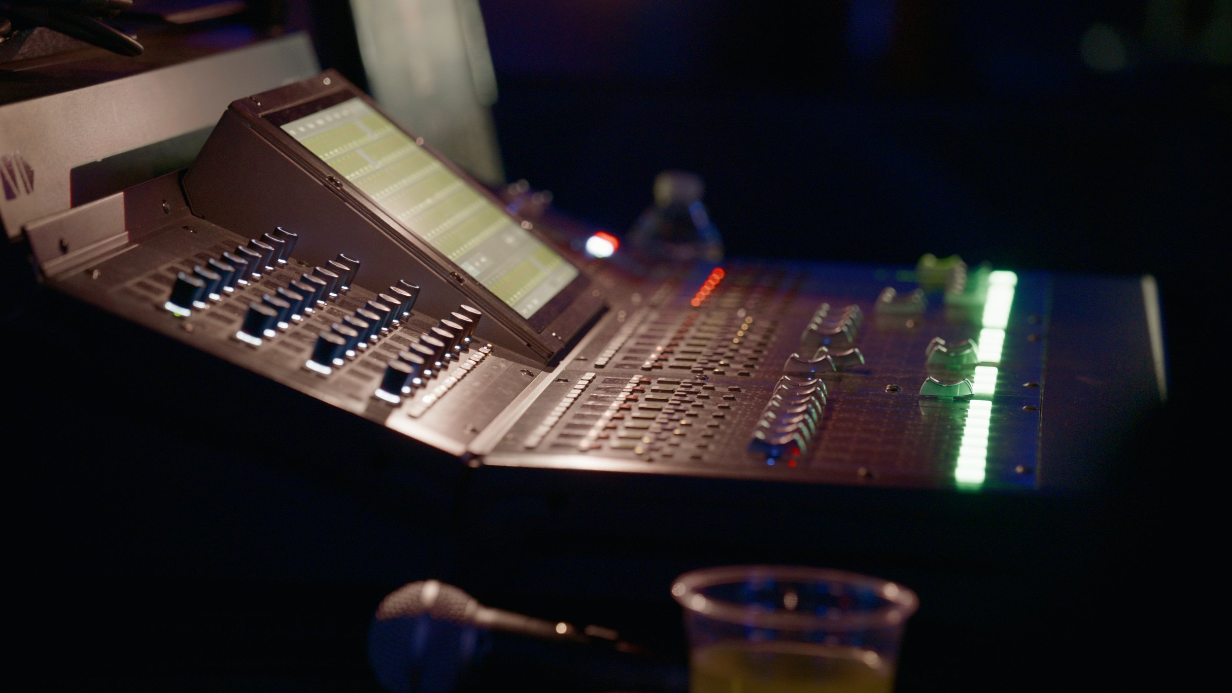 Close-up of a professional audio mixing desk with faders, knobs, and a digital display, illuminated by colored lights in a dark environment.