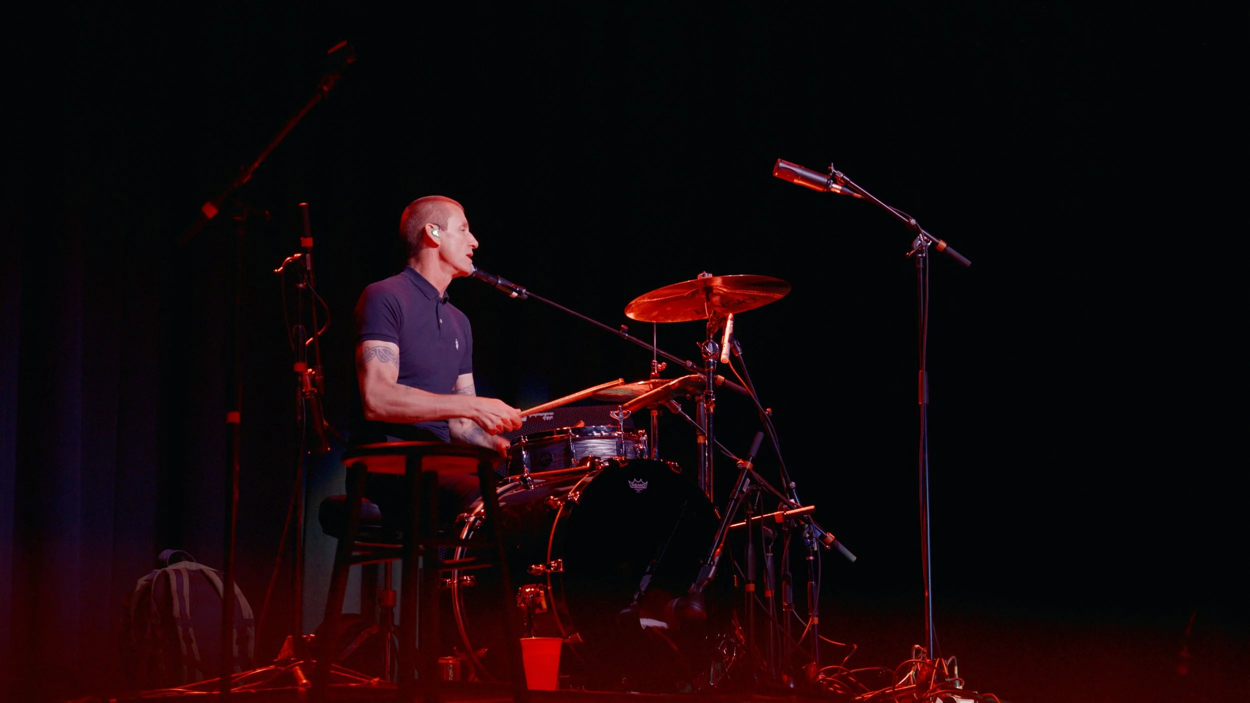 A musician playing drums on a stage with a black background, surrounded by microphones and equipment, illuminated by red stage lighting.