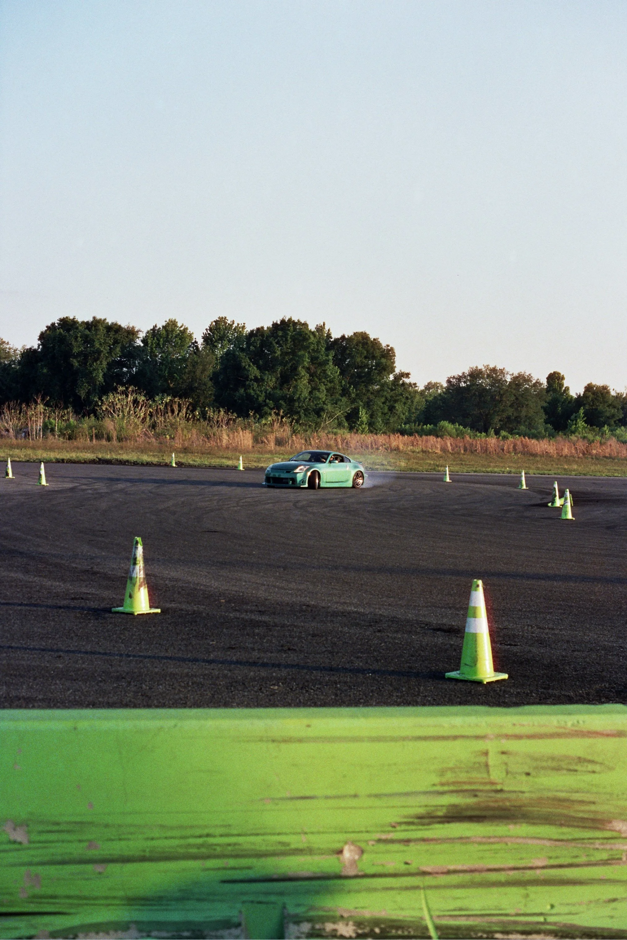 A car drifting around orange and white traffic cones in an open parking lot with a background of trees and sky.