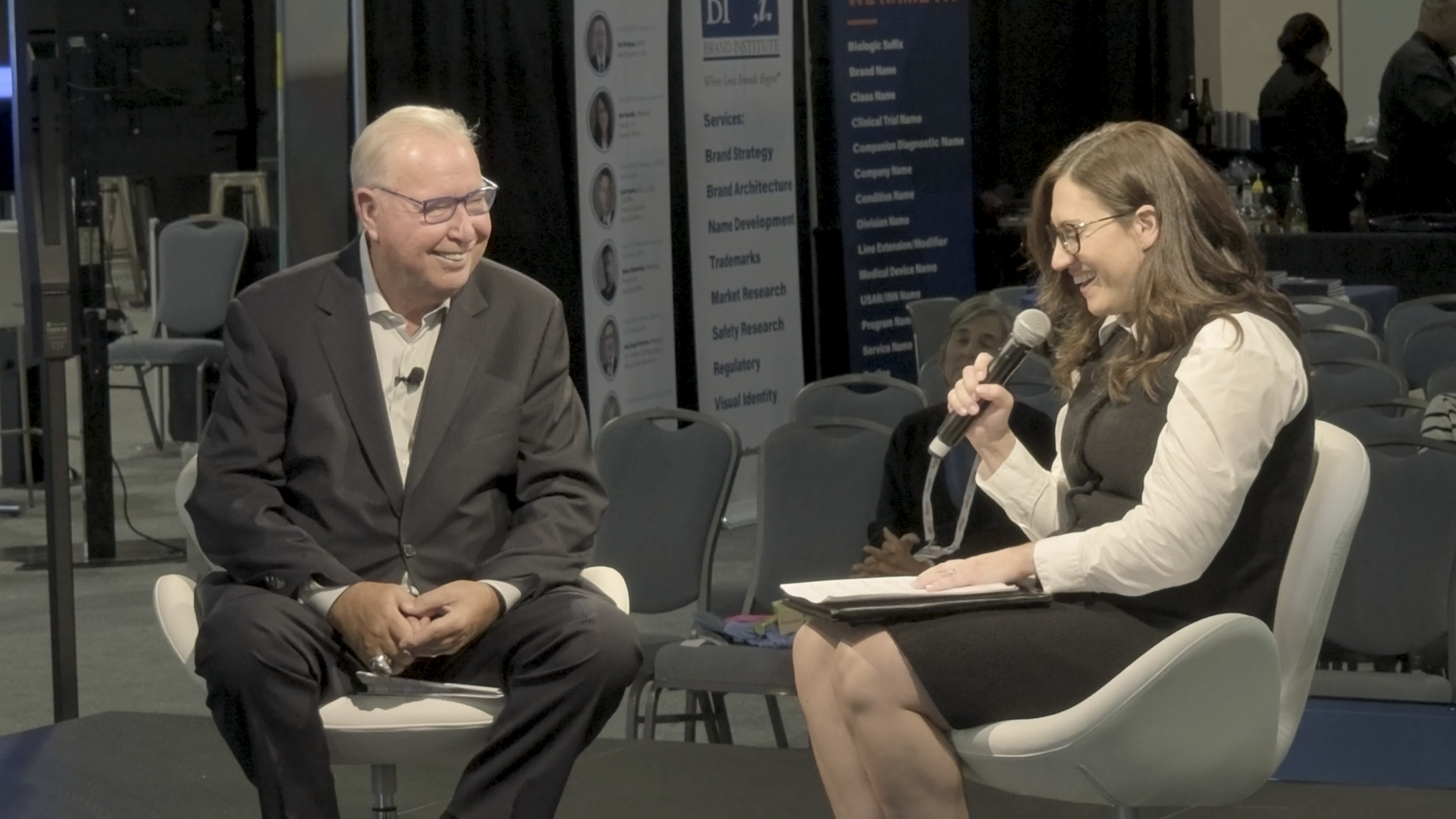 A man in a gray suit and glasses sitting and smiling during an interview or conversation. A woman with glasses, holding a microphone, sitting across from him and smiling. They are in a setting with empty chairs and event banners in the background.