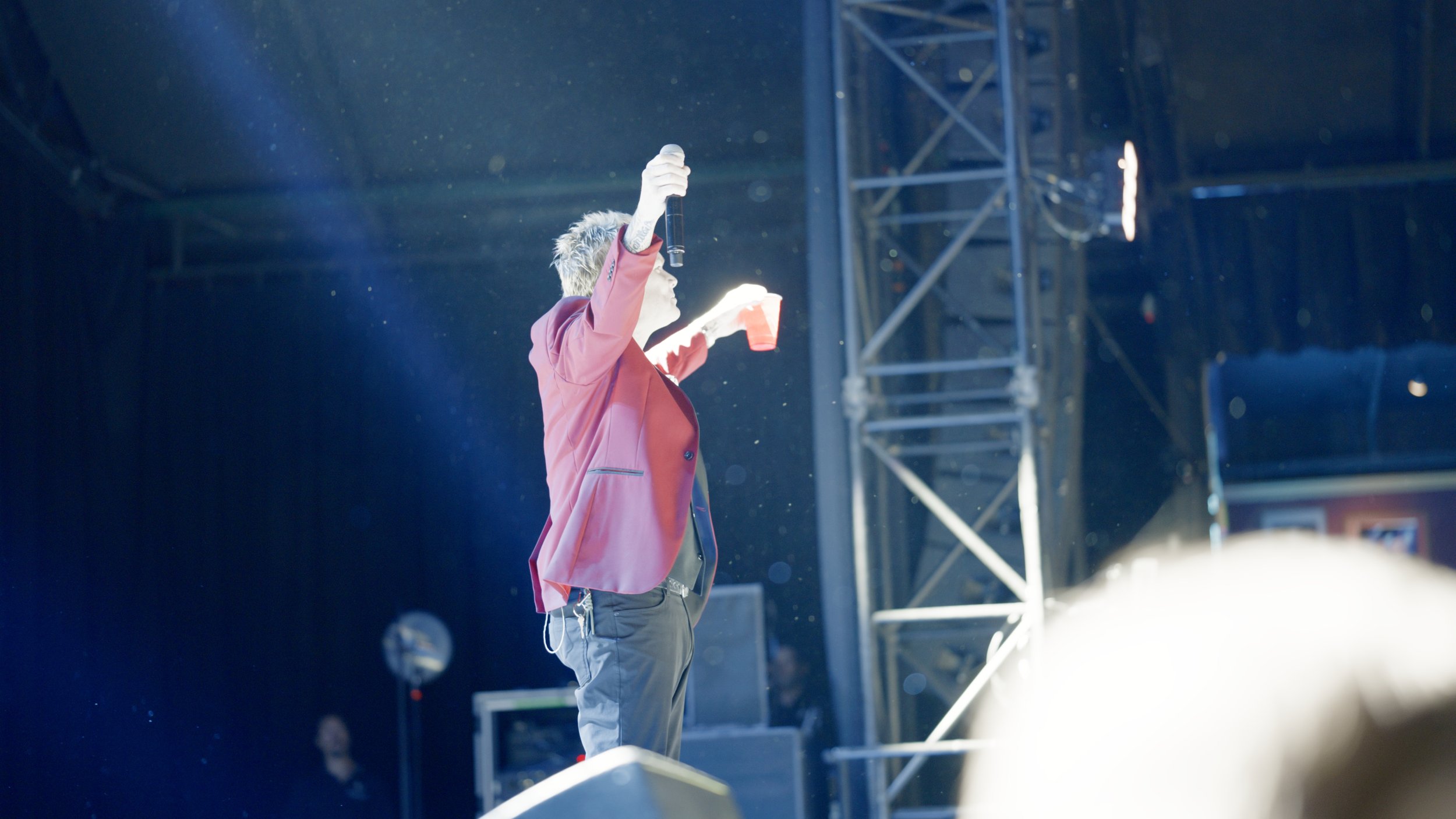 A performer with styled hair and tattoos on arm holding a microphone and a red cup on stage, with a metal scaffolding structure in the background, under stage lighting.