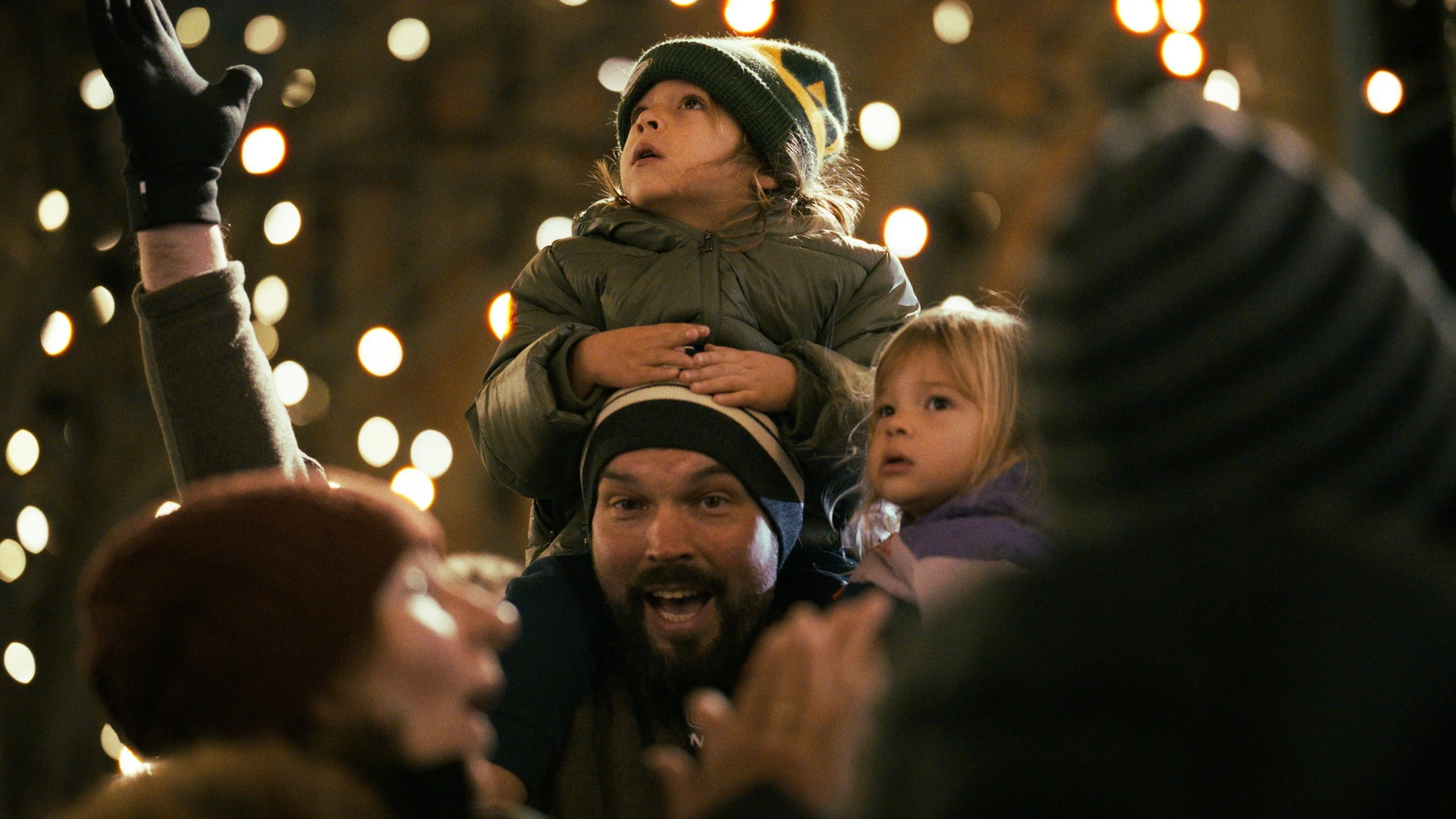 A man with a beard smiling and looking up, carrying two young girls on his shoulders during a nighttime outdoor celebration with string lights in the background.