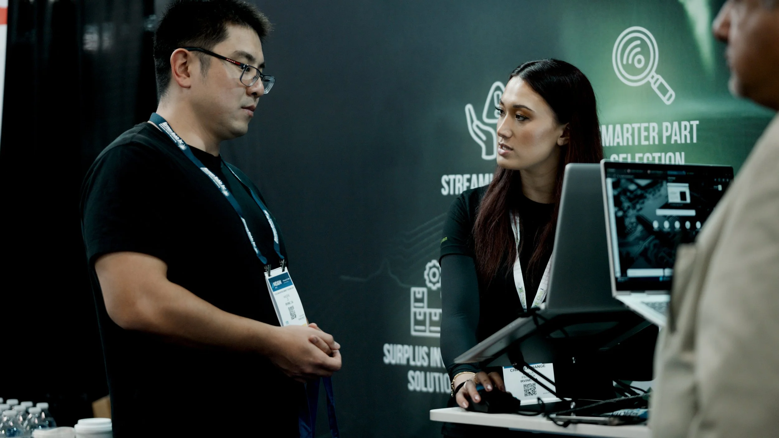 Two men and a woman appear to be engaged in a conversation at a technology booth, with one of the men wearing glasses and holding papers, and the woman working on a laptop with a digital display. The background features icons related to streaming, sm