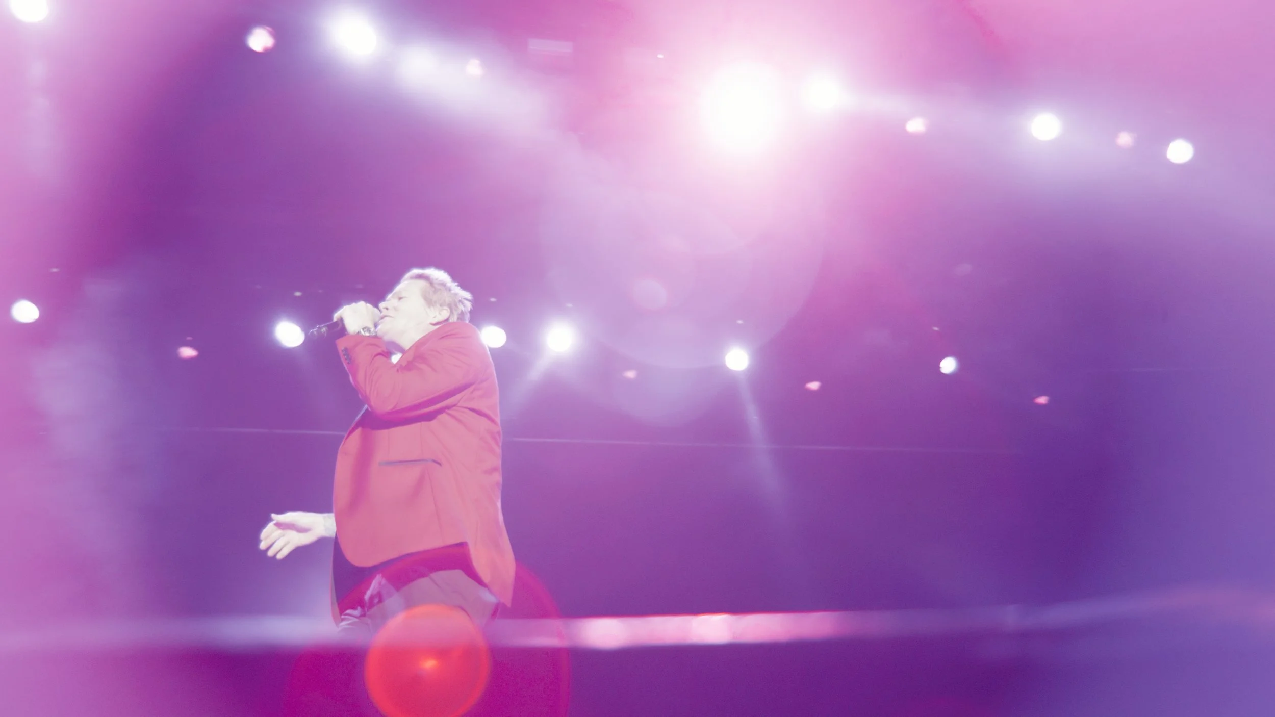 Performer singing on stage with bright purple and pink lights and lens flares.