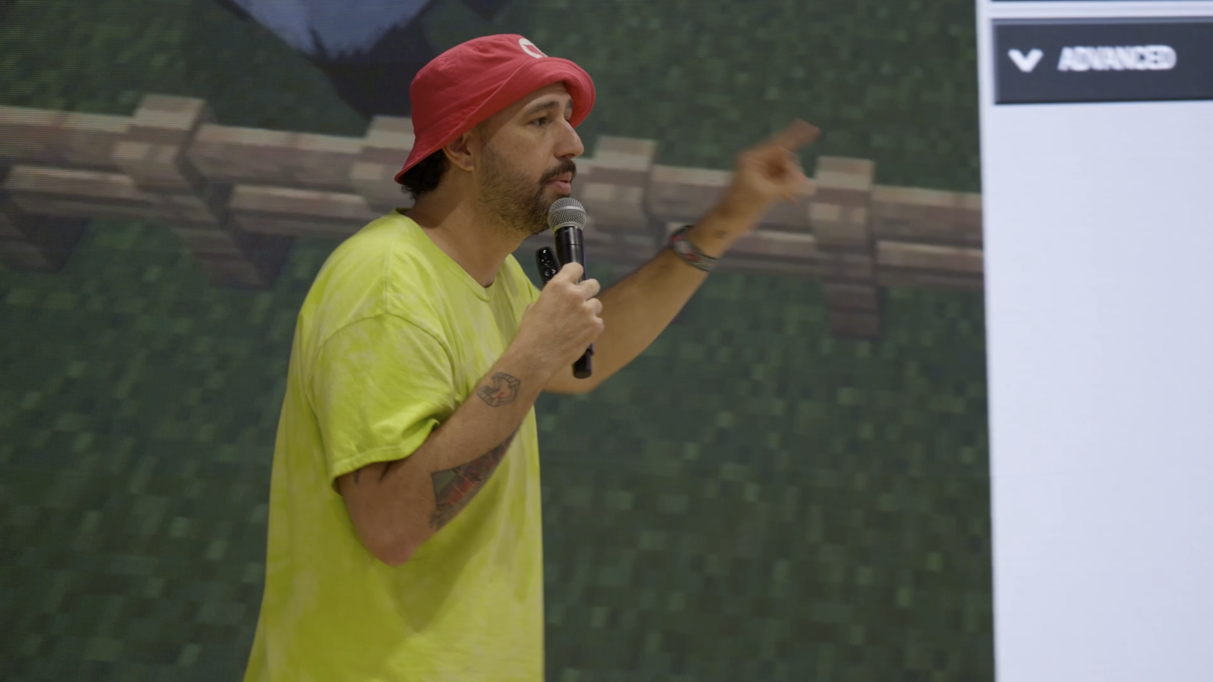 Man with tattoos wearing a yellow shirt and red bucket hat, holding a microphone, giving a presentation in front of a digital screen.