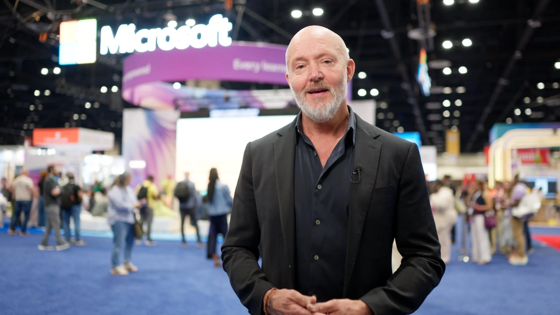 A man with a bald head and gray beard wearing a black suit jacket and shirt stands at a technology conference or expo with a crowded background and large Microsoft signs.