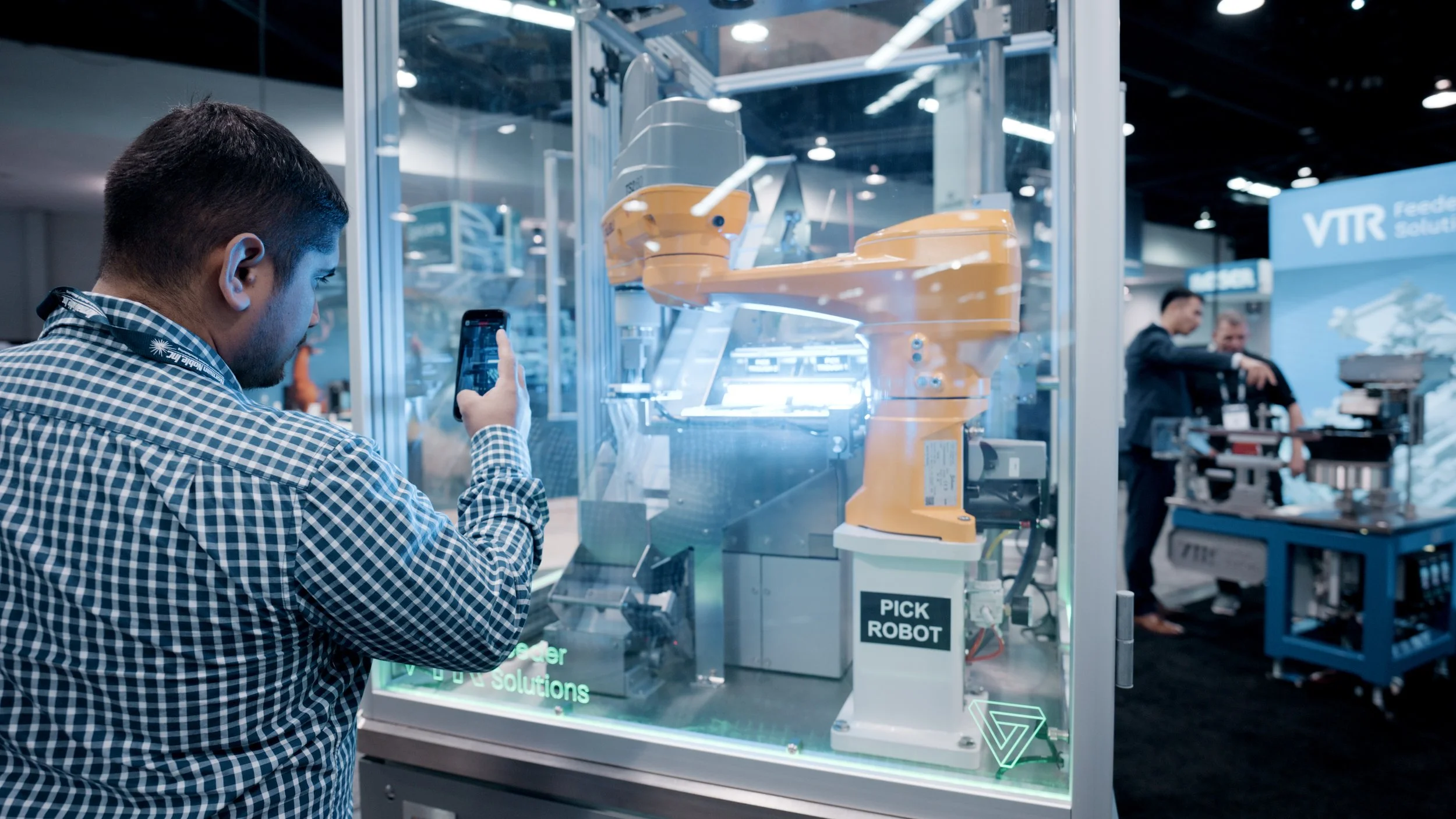 A man with a checkered shirt taking a photo of an orange industrial robot arm inside a glass enclosure at a technology expo.