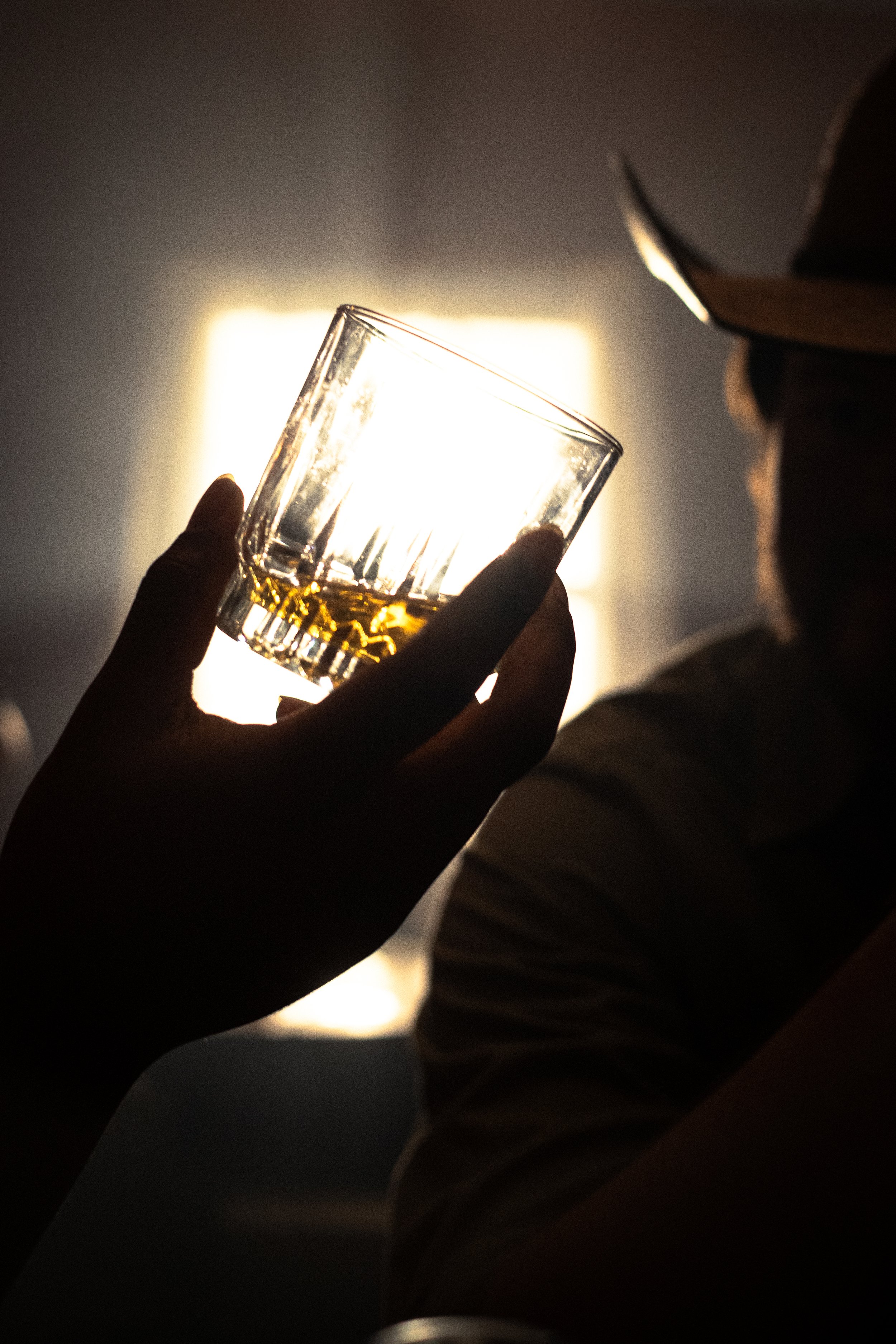 A person holding a glass of whiskey in front of a bright light, with another person visible in the background wearing a hat.