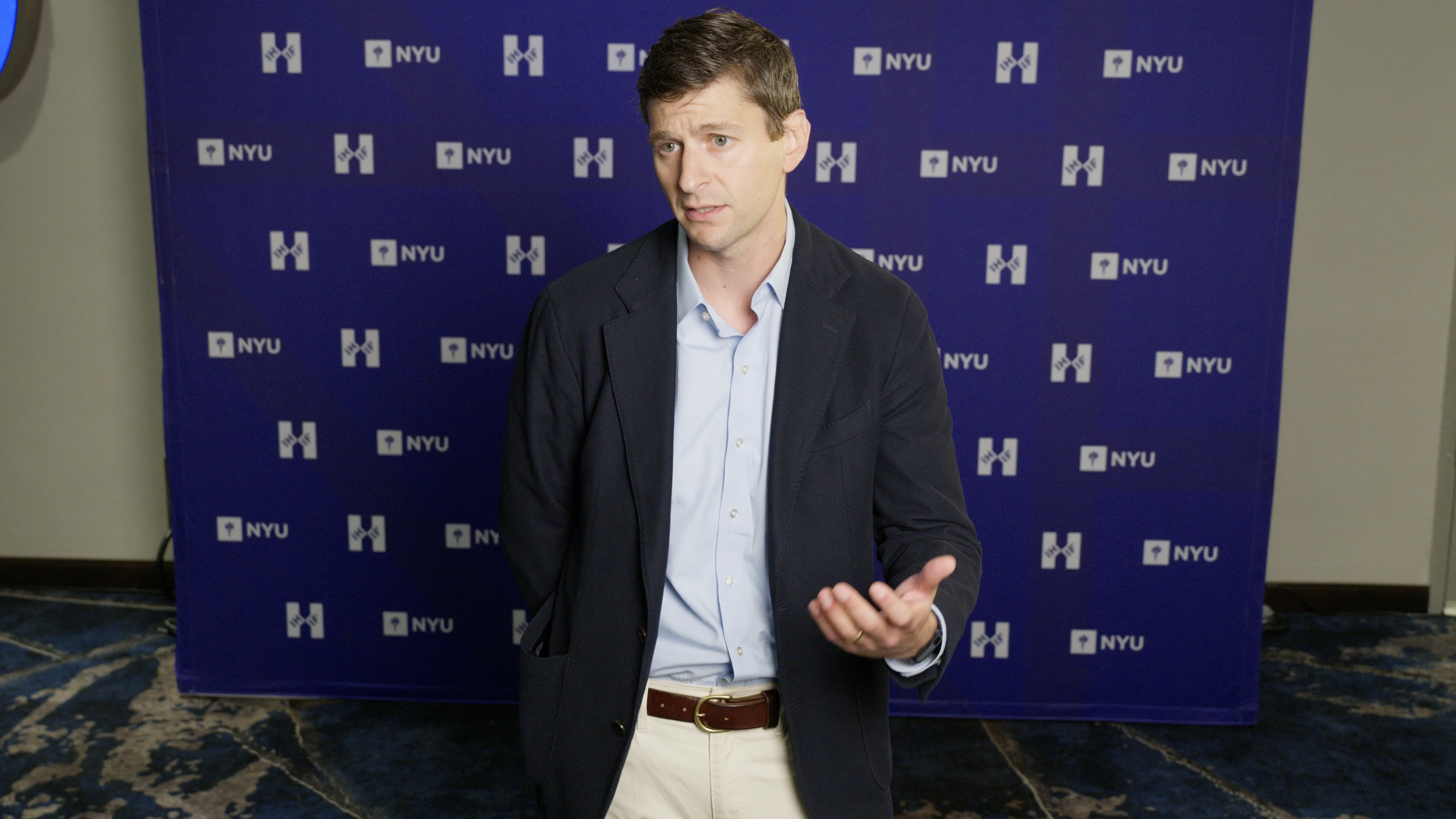 A man in a business jacket and light blue shirt gesturing with his hand, in front of a blue backdrop with the NYU logo repeated on it.