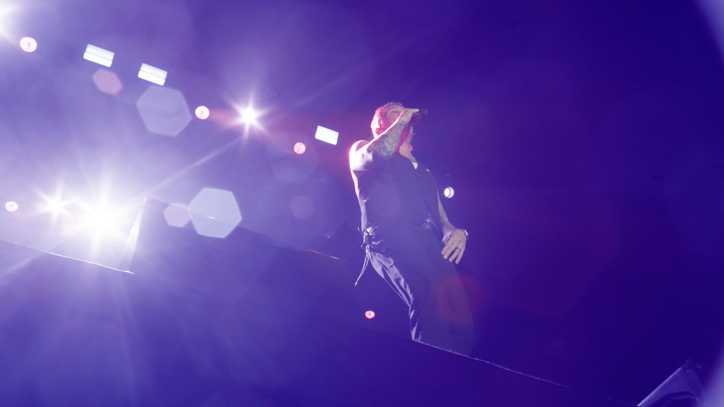 A performer singing on stage with purple lighting, wearing a black outfit, visible tattoos, and holding a microphone.