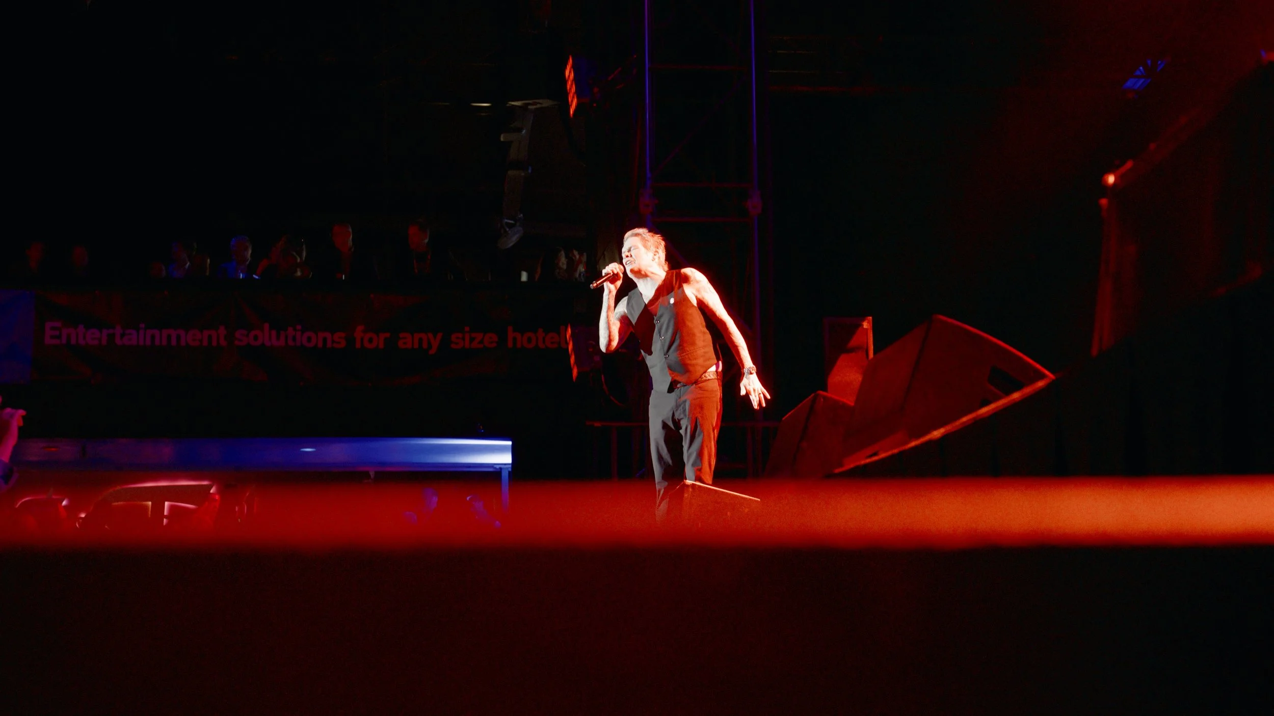 Male performer singing with a microphone on stage, illuminated by red lighting, with an audience and a sign reading 'Entertainment solutions for any size hotel' in the background.