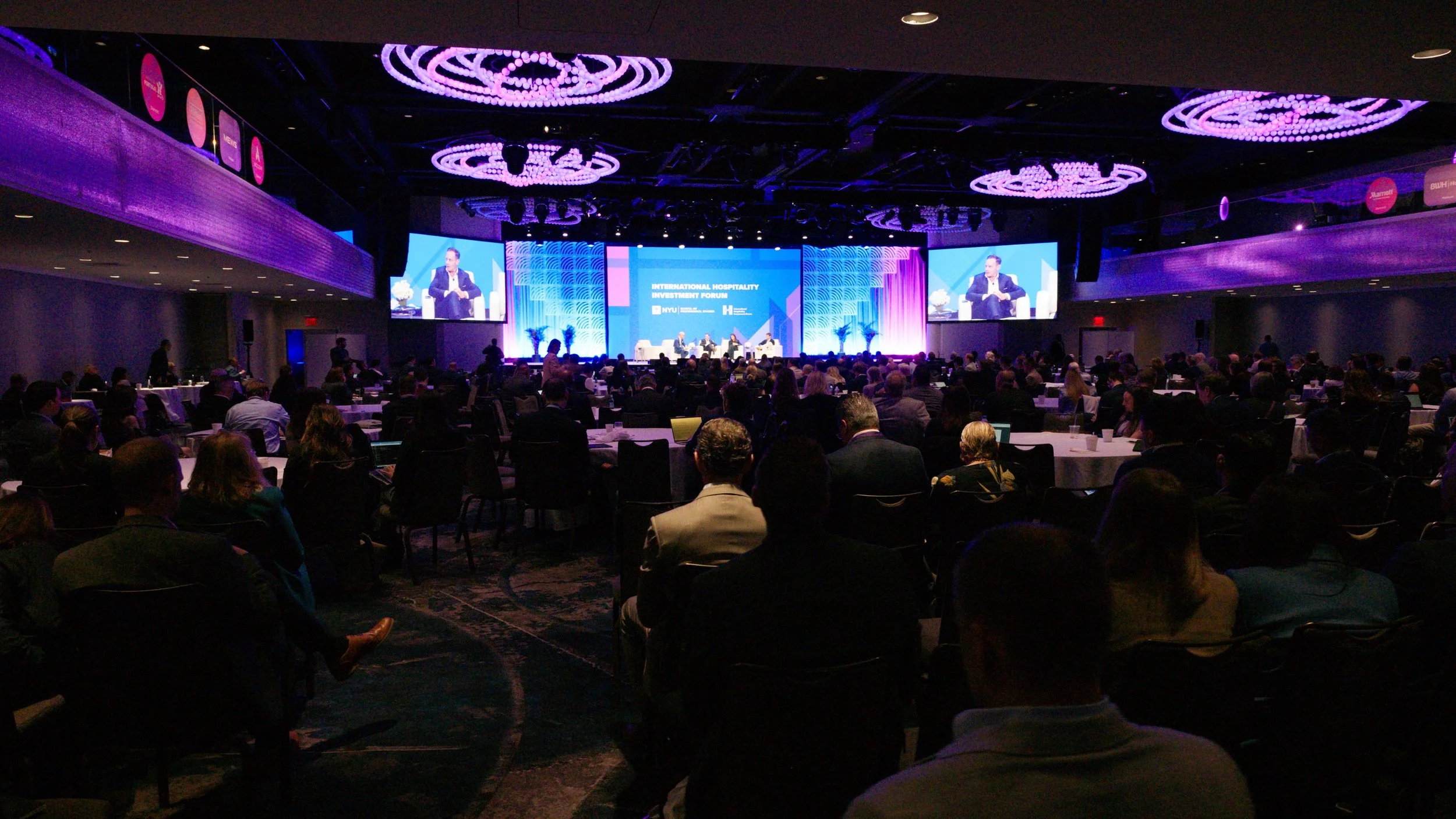A large conference room filled with attendees facing a stage with a panel of speakers and a large screen displaying the event name, 'International Hospitality Investment Forum'.