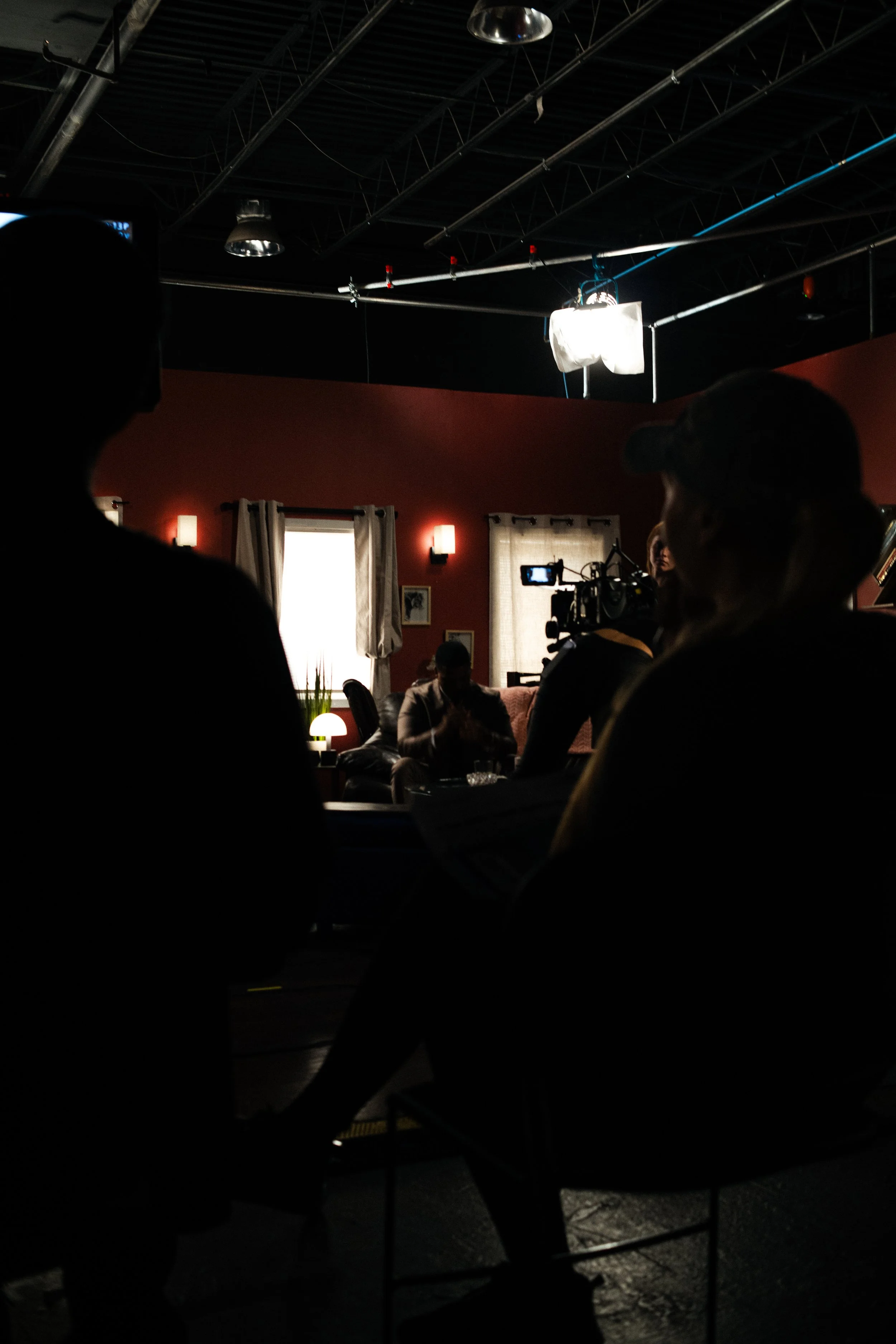 Inside a dimly lit room with red walls, several people are seated, possibly watching a filming or photoshoot. A person is operating a camera aimed at a scene or person, and there are visible professional lighting and equipment overhead. Windows with 