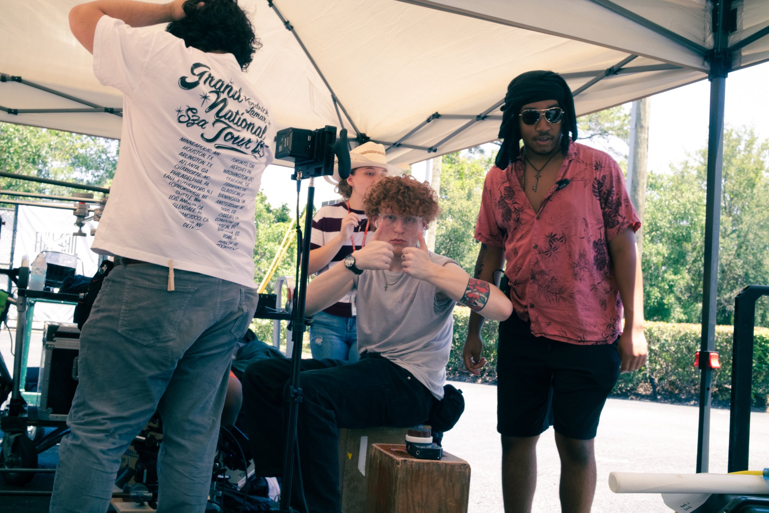 A group of four people under a white canopy tent outdoors. One person with curly hair and glasses is sitting on a chair, giving two thumbs up. Two others are standing nearby, one wearing sunglasses and a pink Hawaiian shirt, and the other in a stripe