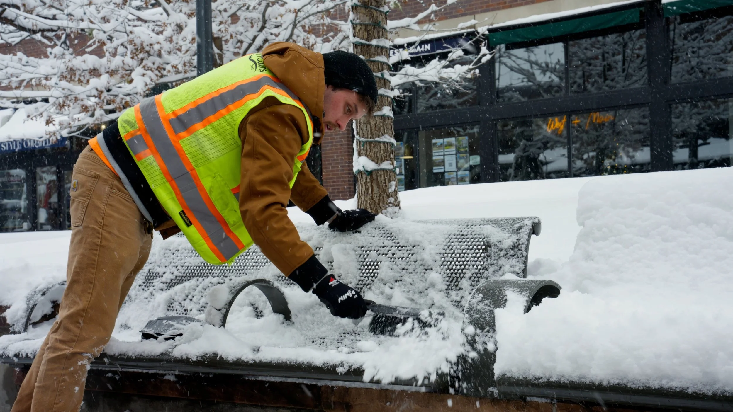 A man in a yellow safety vest clearing snow from a bench during winter.