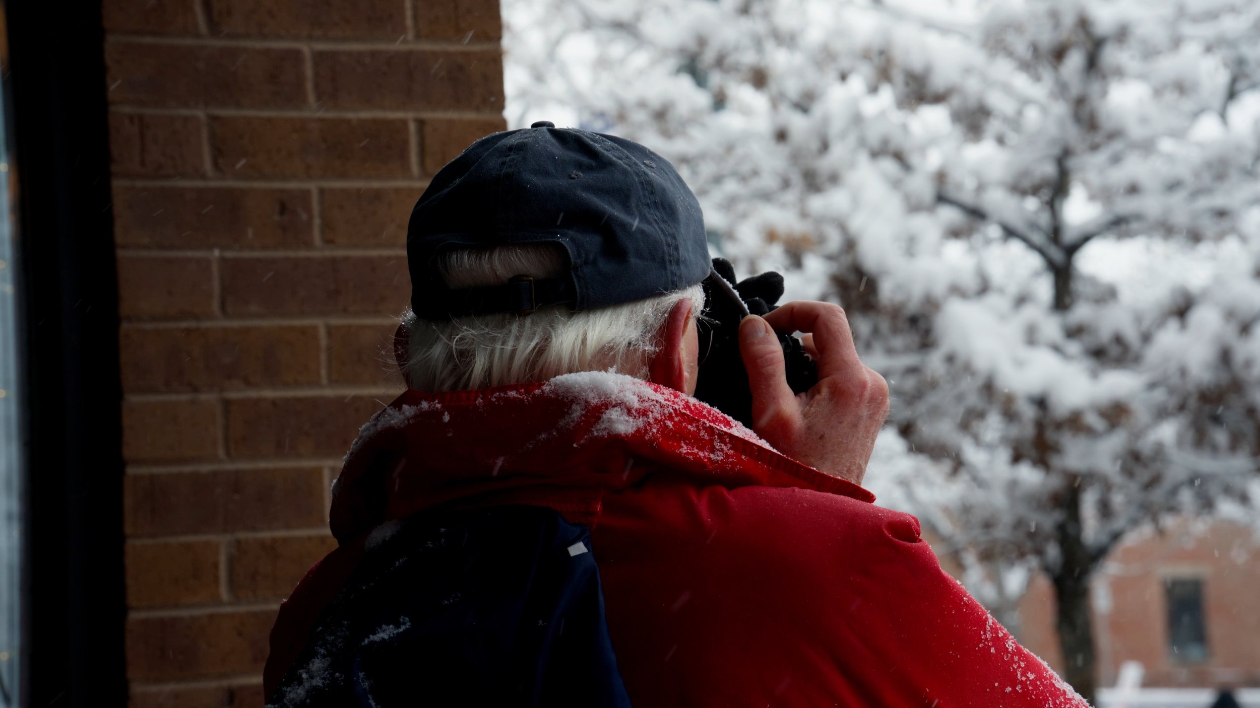 Person with white hair, wearing a dark baseball cap and red jacket, taking a photograph outside in snowy weather.