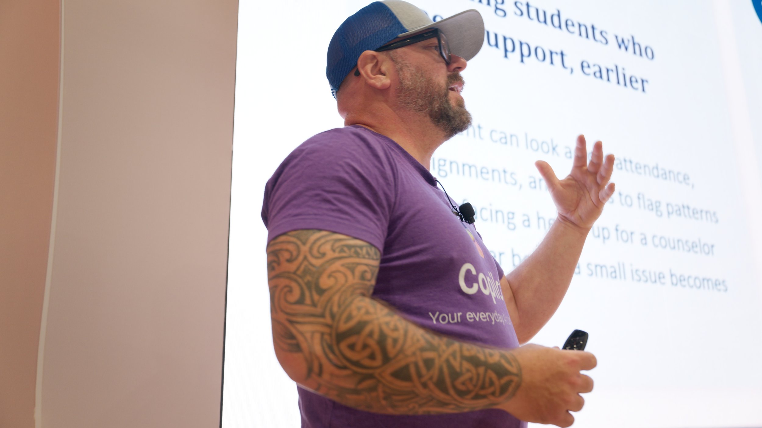 A man wearing a baseball cap, glasses, and a purple T-shirt giving a presentation in front of a screen displaying text. He has tattoos on his arms and holds a remote control in one hand.