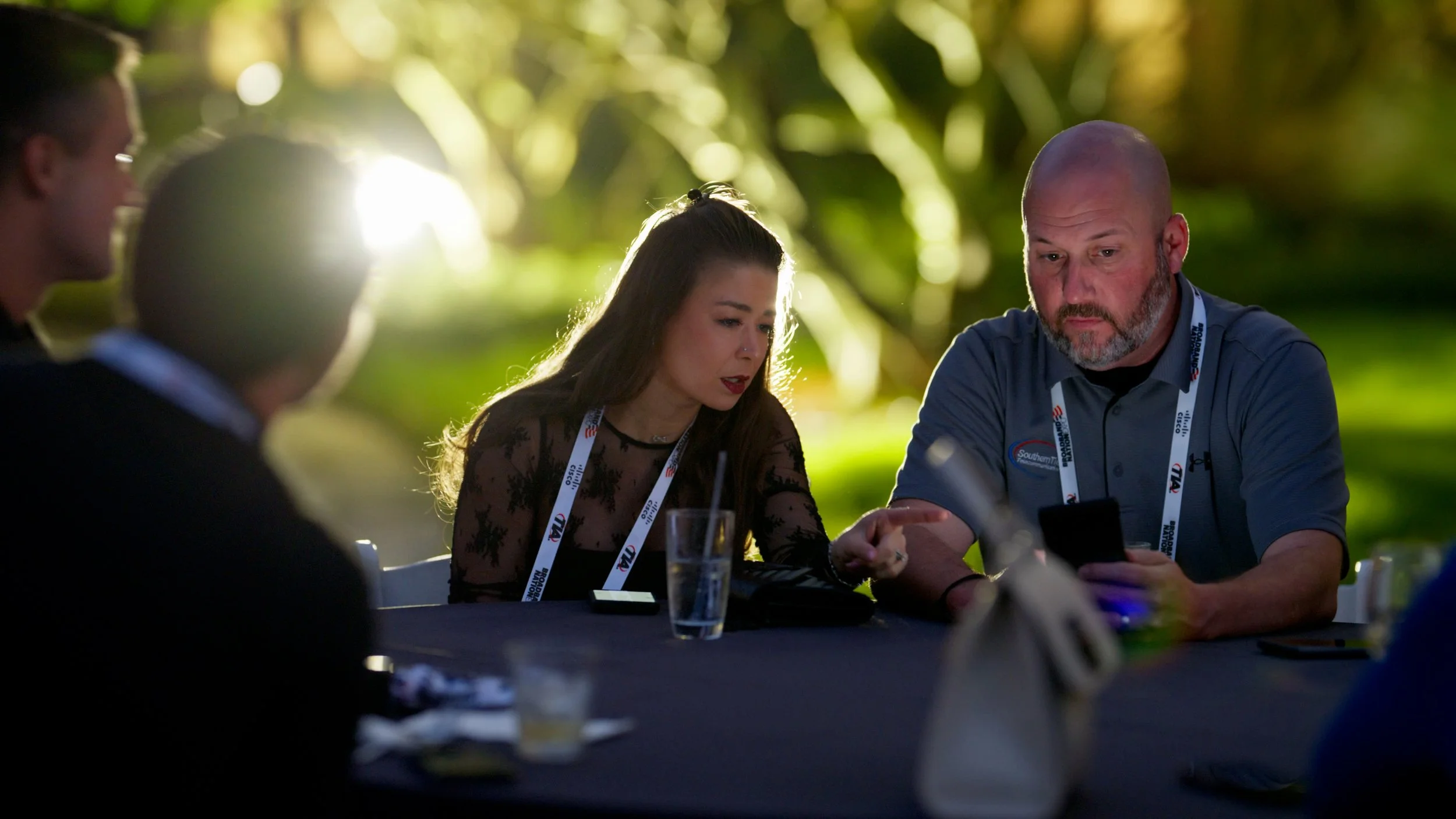 Group of four people seated at a table outdoors during daytime, looking at their phones, with a bright sunlight in the background and green foliage behind them.