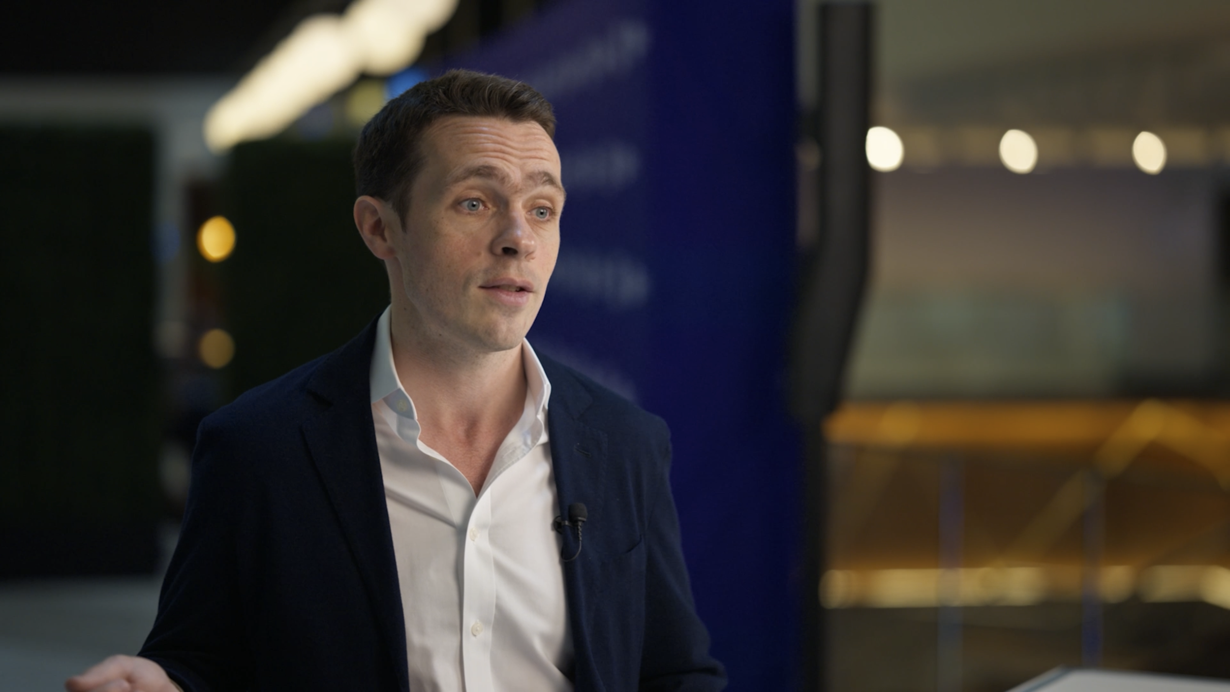 A man in a dark jacket and white shirt speaking or being interviewed indoors, with a blurred background of lights and modern decor.