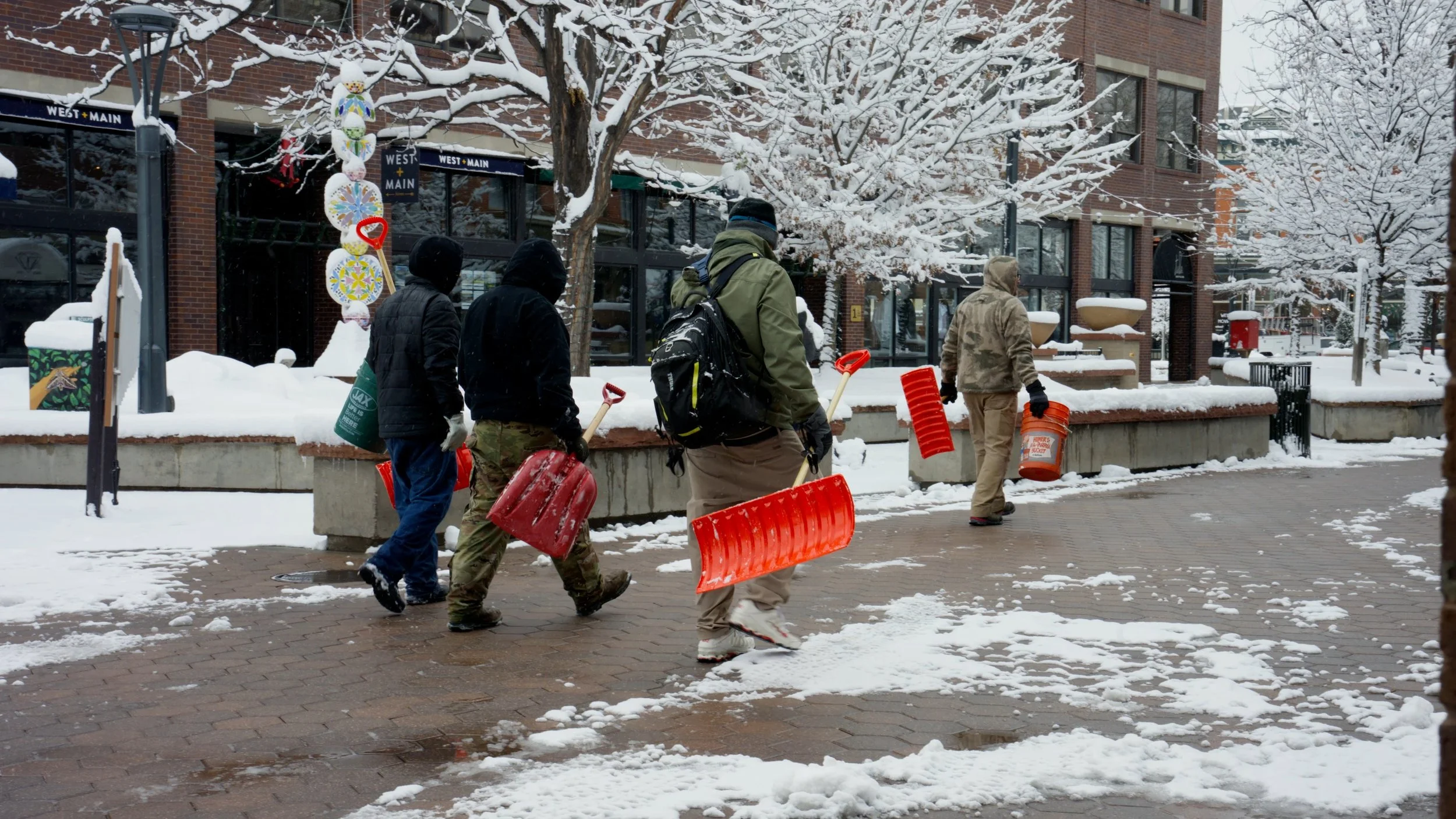 Five people walking on a snowy sidewalk carrying snow shovels and orange snow rakes, with a brick building and snow-covered trees in the background.