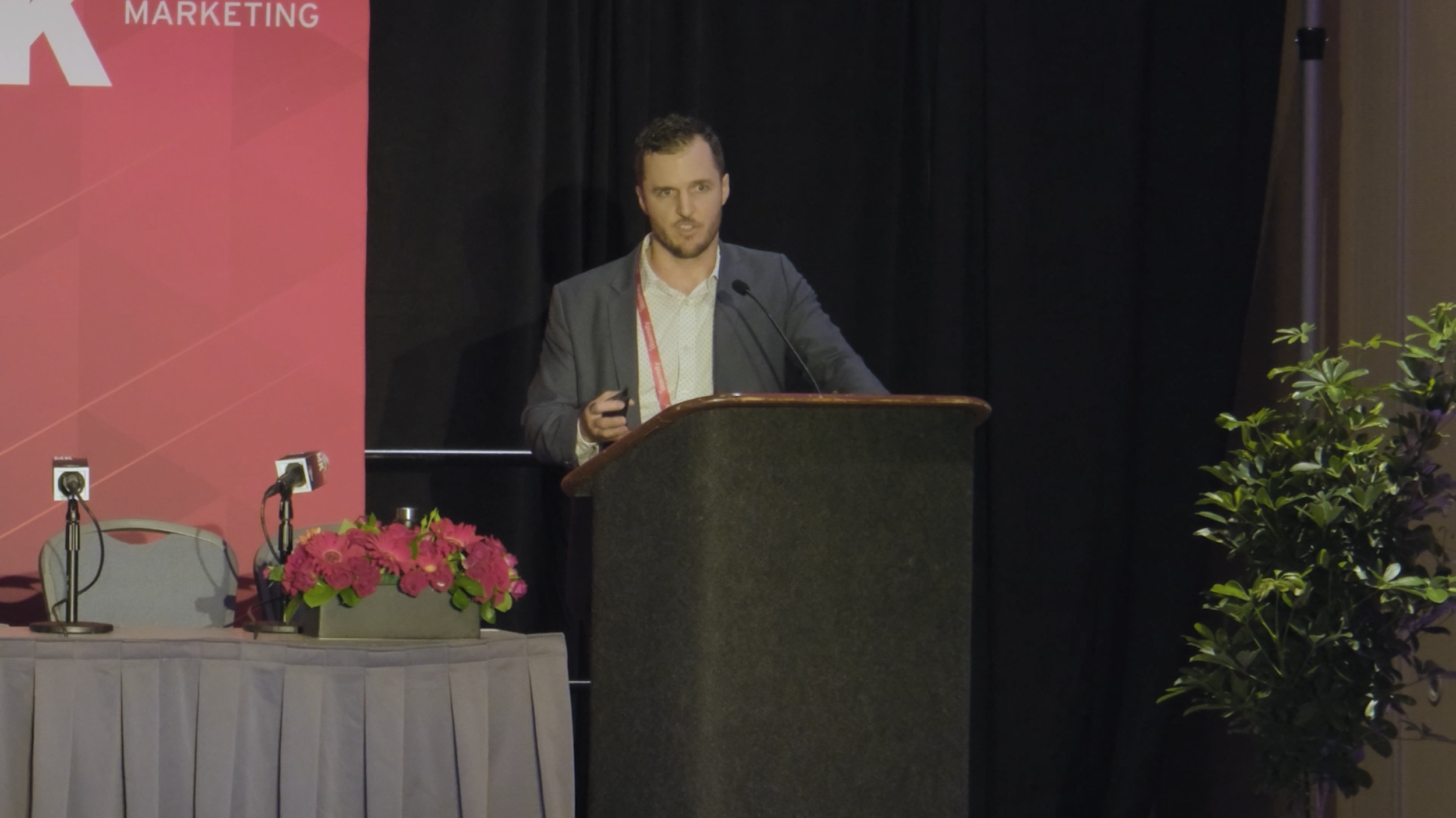 A man in a gray jacket and white shirt standing behind a podium giving a presentation at a conference with a black curtain background. There are pink flowers on the table in front of him and multiple microphones on the table. The left side has a pink and red banner with the word 'MARKETING' on it.