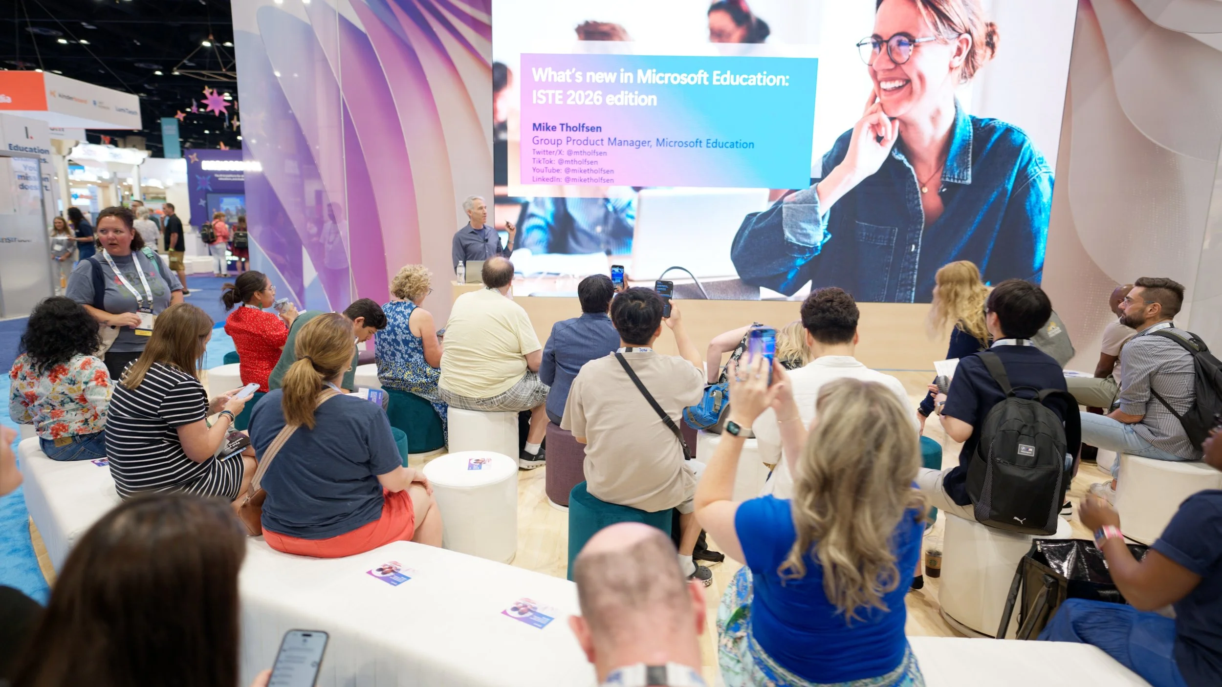 A speaker at a conference presenting on Microsoft Education, with a large screen behind them displaying the topic and the presenter's name, Mike Tholfsen. The audience, sitting on chairs and stools, listens attentively, some taking notes or photos, i