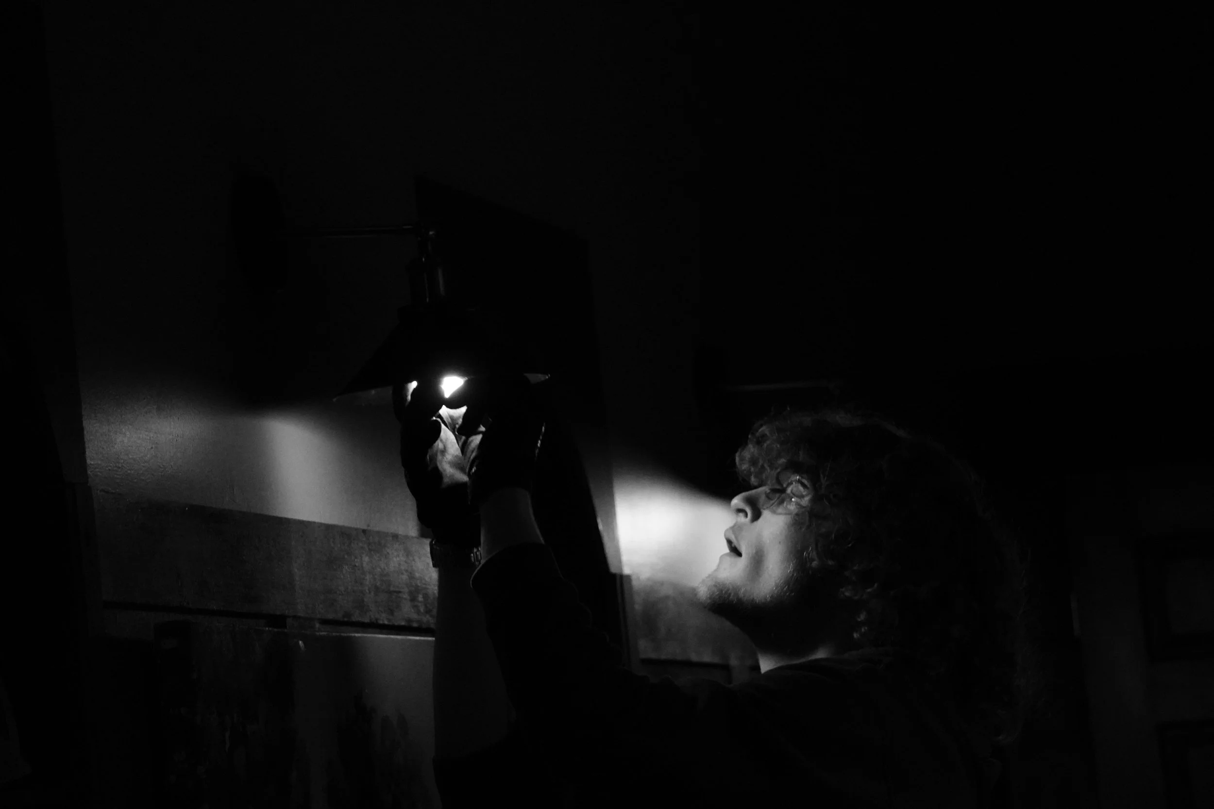 A person with curly hair, wearing glasses, is looking at a small object held in their hand that emits light, in a dark room with minimal lighting.