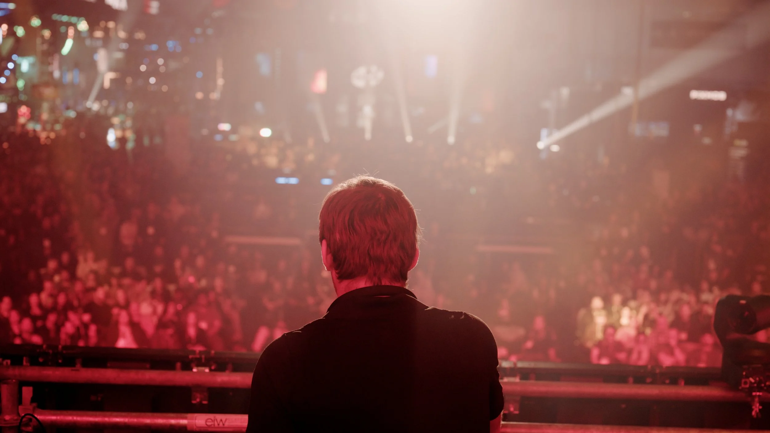 A person with red hair seen from behind on a stage, facing a large crowd in a concert venue with bright stage lights and a cityscape backdrop.